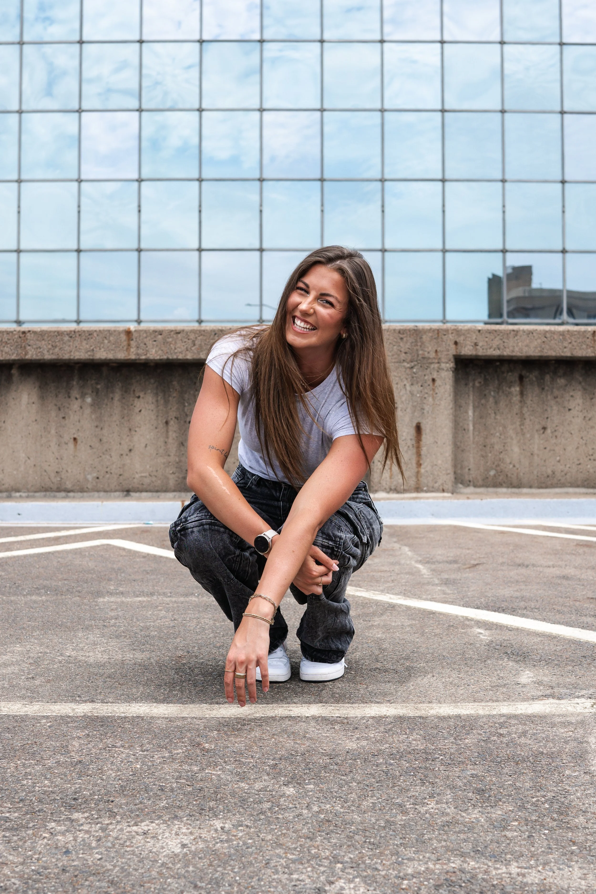 A young woman with long brown hair squats on a parking lot pavement in front of a concrete barrier and a large glass window reflecting a cloudy sky. She is smiling, wearing a white T-shirt, dark patterned pants, white sneakers, and a wristwatch, with