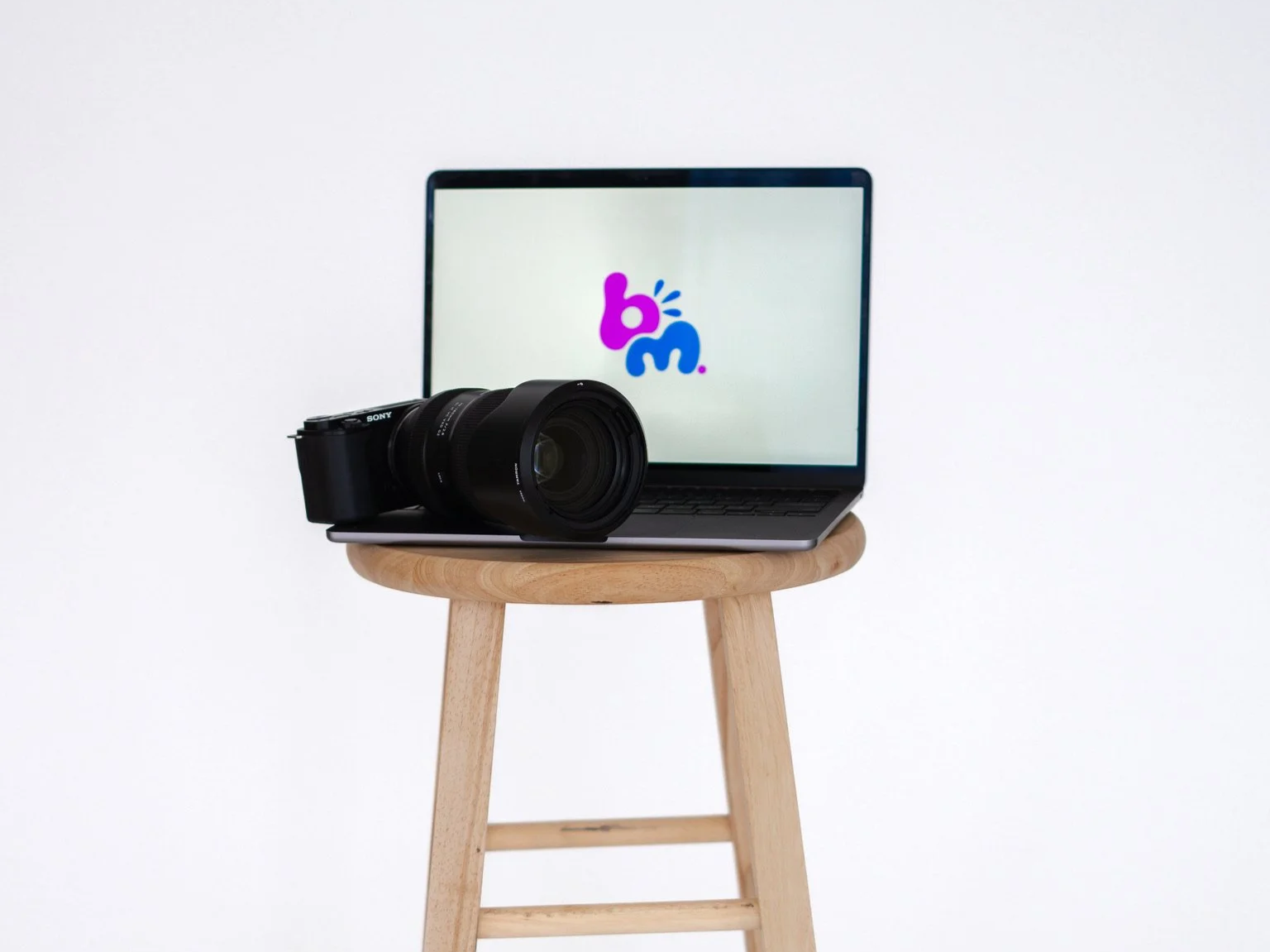 A wooden stool with a laptop and a camera on top, the laptop screen displays a logo with colorful letters 'b' and 'm'