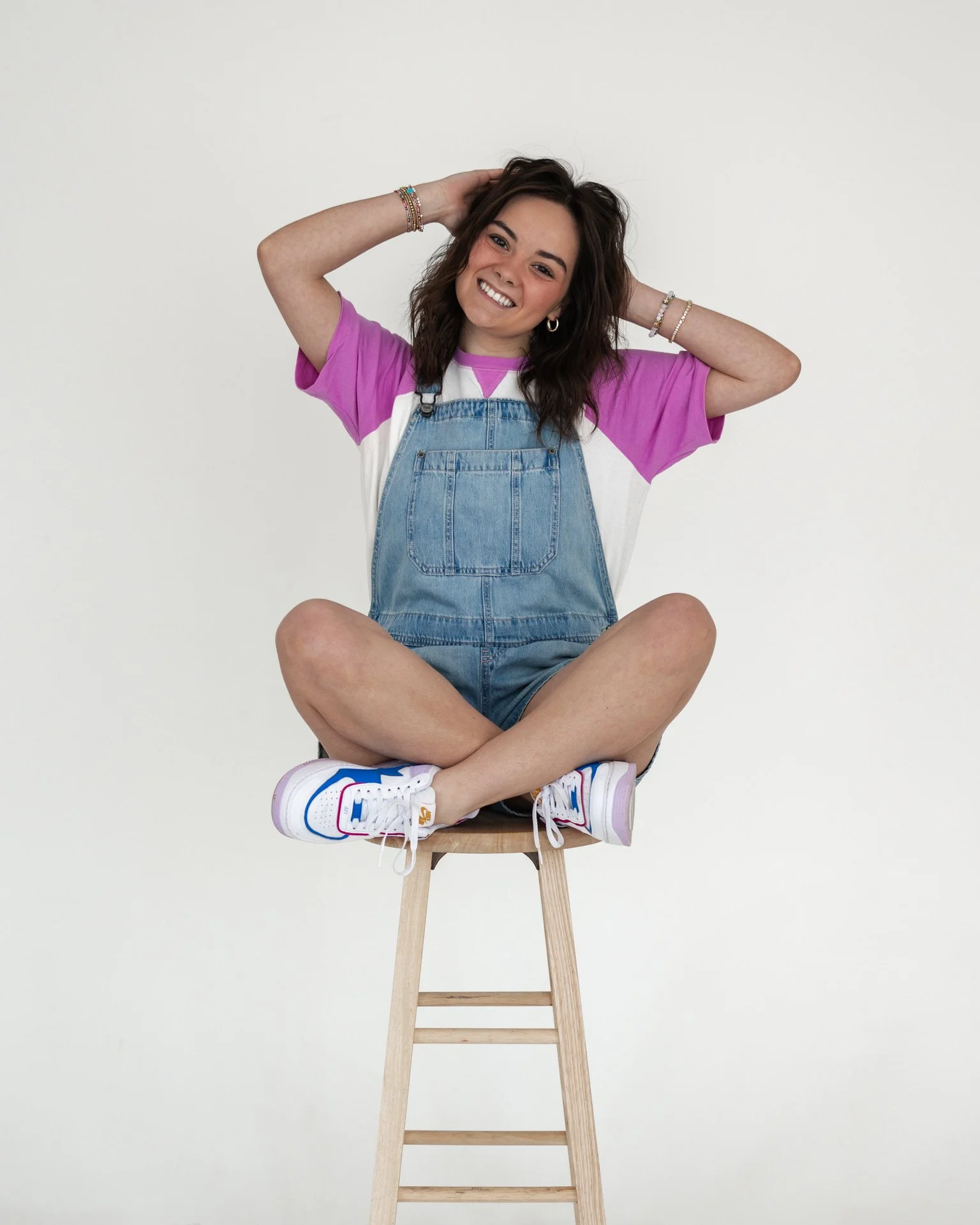 A young woman sitting cross-legged on a wooden stool, smiling, with her hands behind her head. She is wearing a pink and white t-shirt, denim overalls, colorful jewelry, and white sneakers.