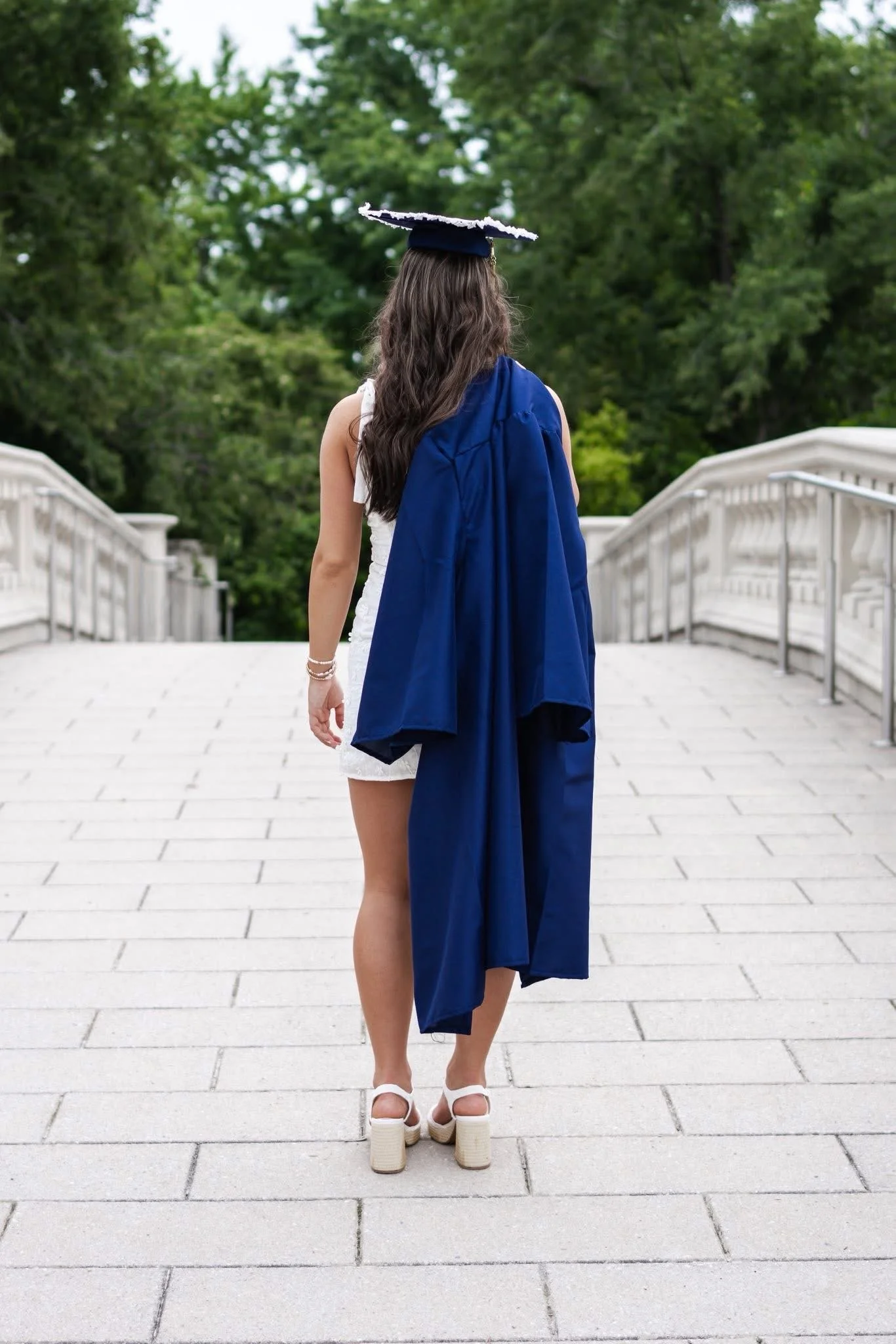 A woman with long wavy brown hair is walking away from the camera on a stone pathway, wearing a graduation cap and gown, white shoes, and a white dress, with a background of green leafy trees.