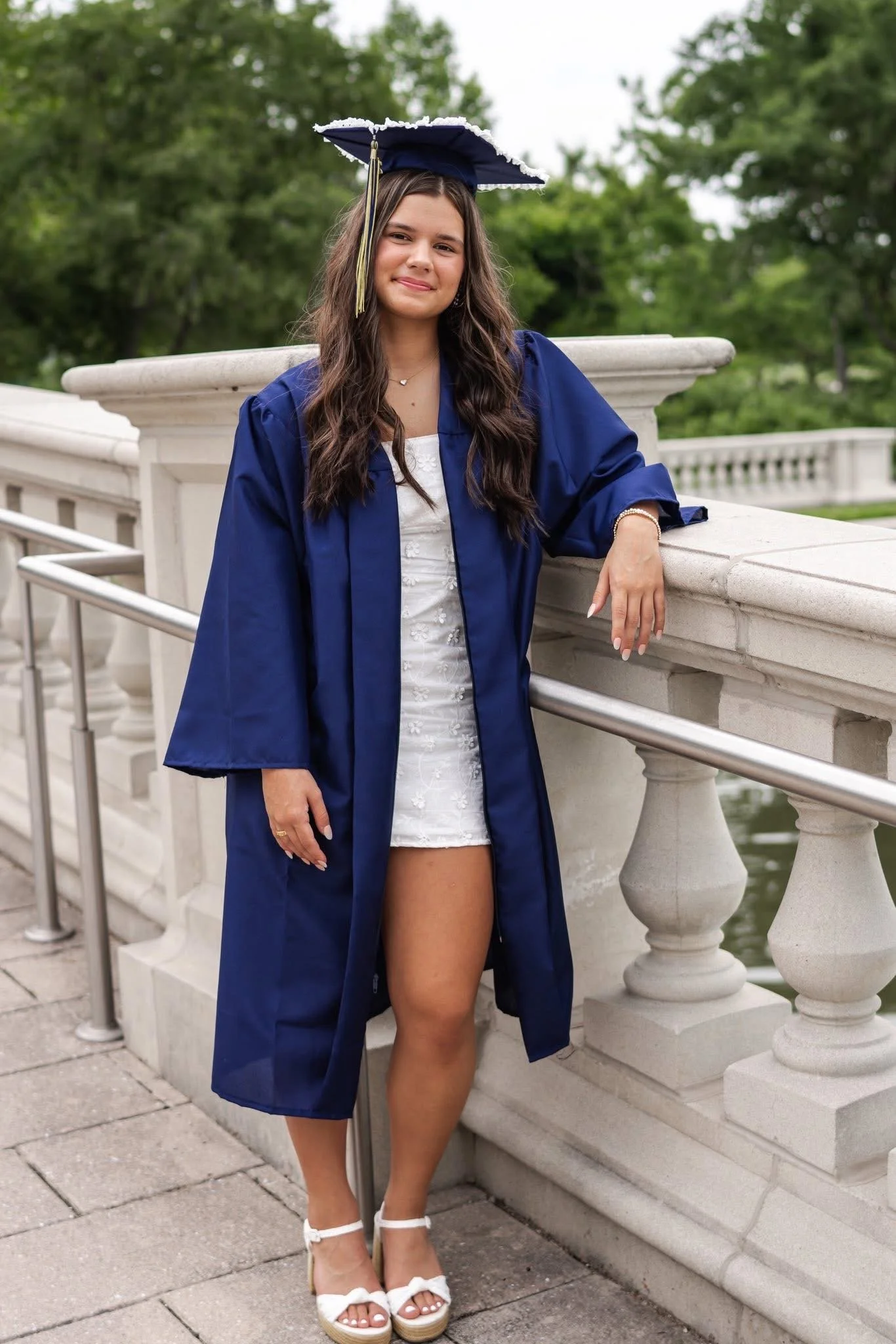 Young woman in a graduation cap and gown standing outdoors near a white stone railing, smiling with greenery in the background.