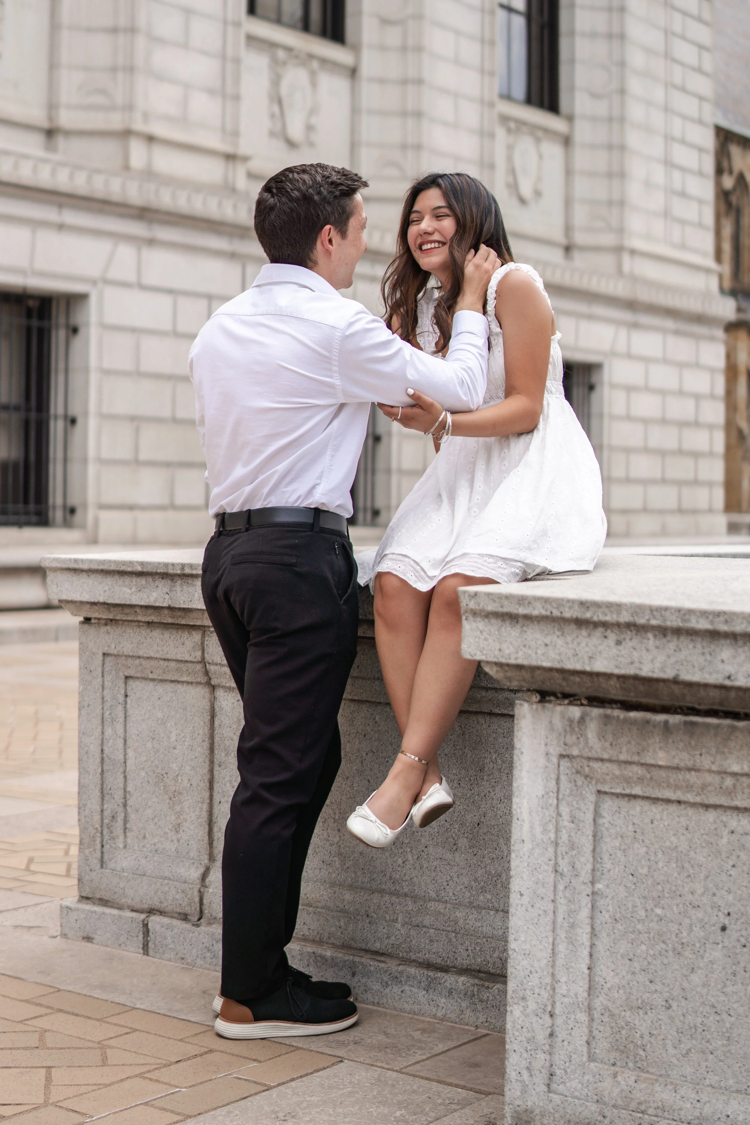 A young man and woman are sharing a joyful moment outdoors. The woman is sitting on a stone ledge, smiling and looking at the man who is standing in front of her. He is gently touching her face and holding her hand. The woman is wearing a white dress