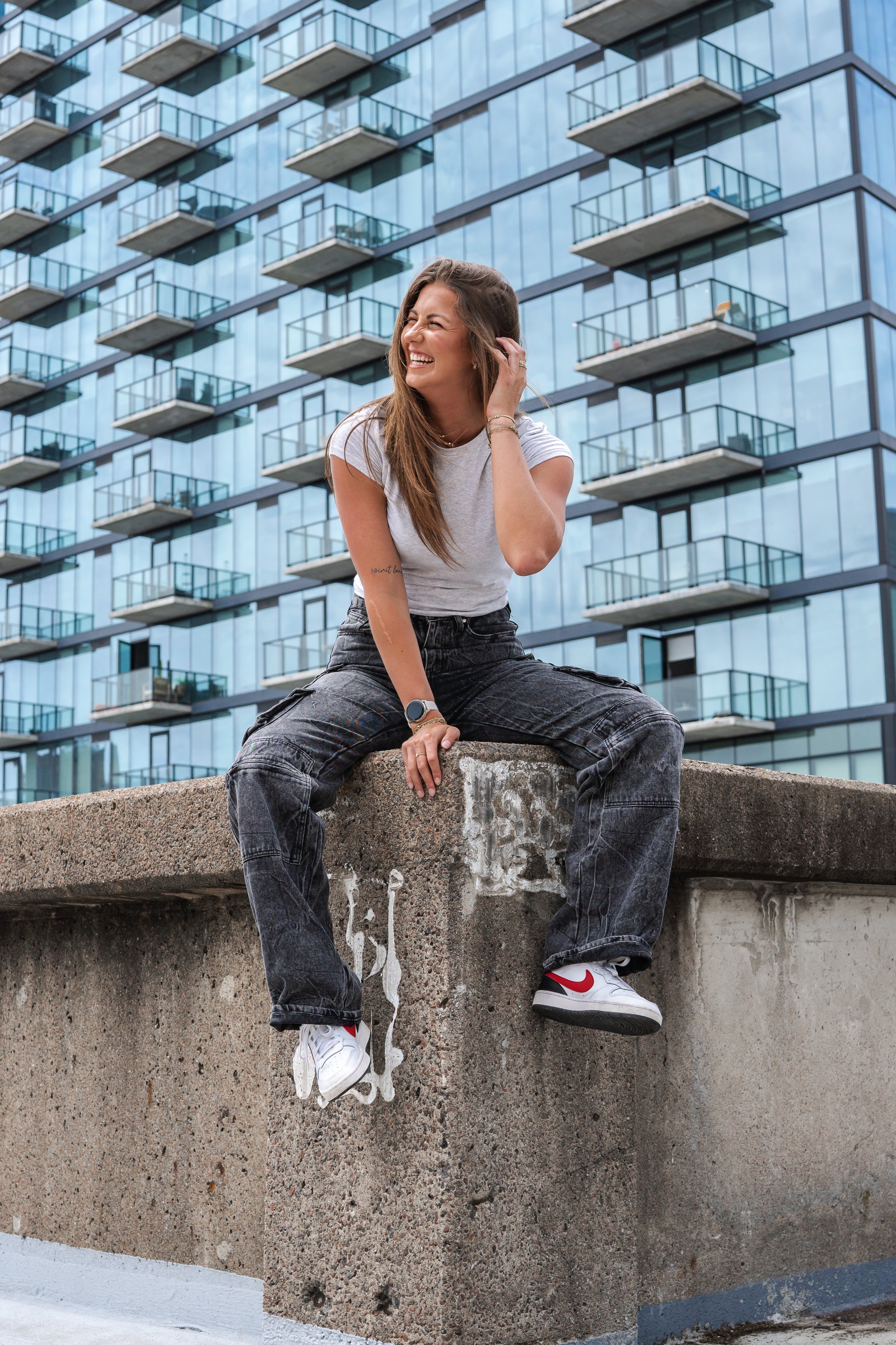 A woman sitting on a concrete ledge in front of a modern glass building, smiling and looking to her left, wearing casual clothes including white and red sneakers.