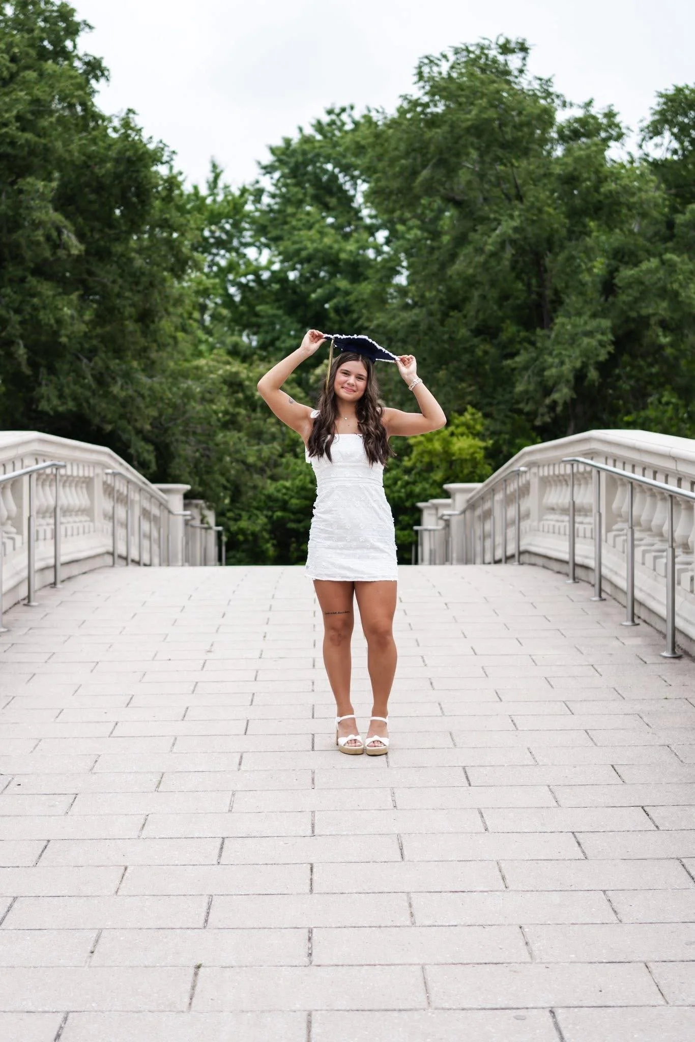 A woman in a white dress holding a graduation cap stands on a stone walkway bridge with greenery in the background.