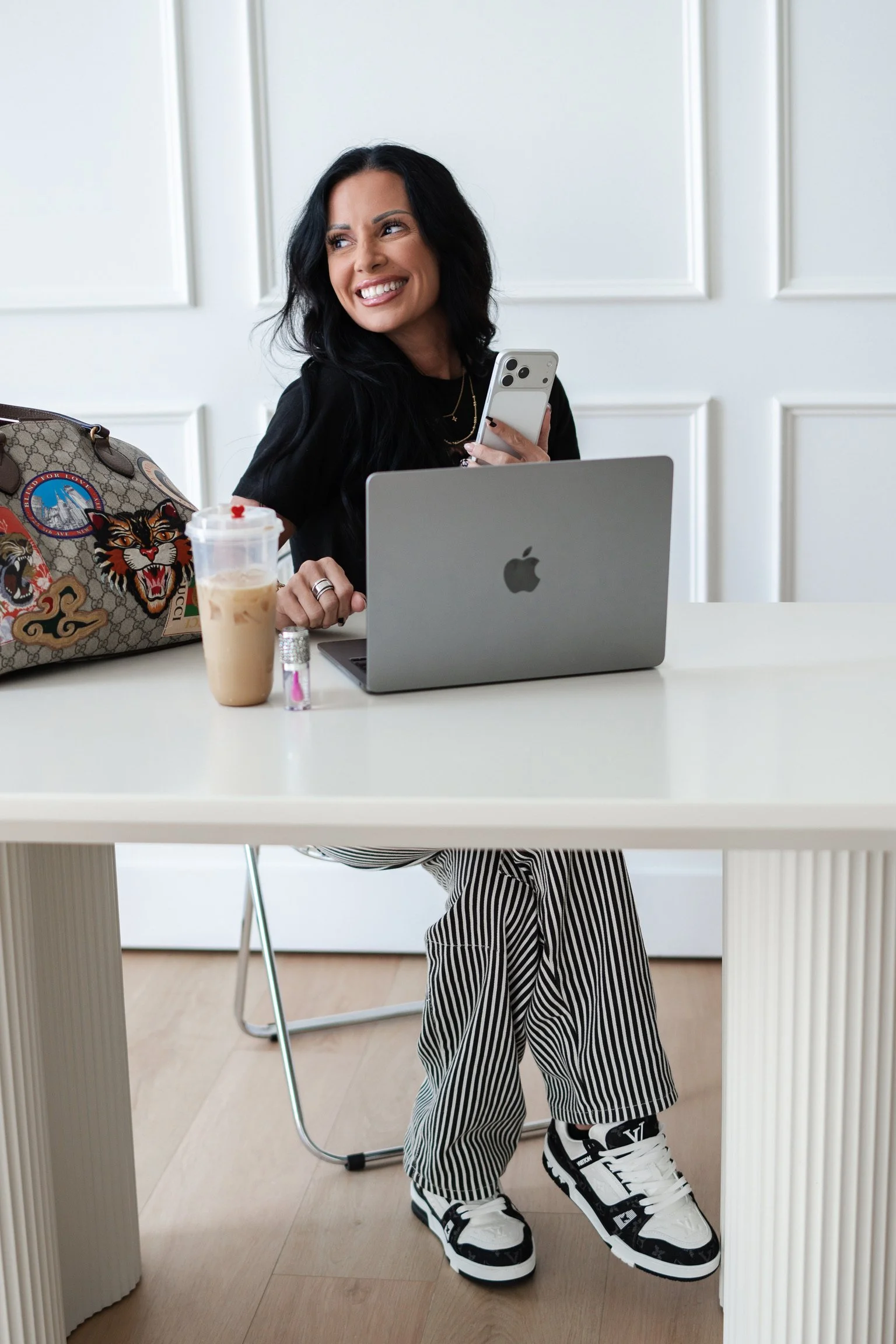 A woman with black hair smiling while sitting at a white table with a silver MacBook, holding a smartphone, wearing black and white striped pants and white sneakers, with a designer bag, iced coffee, and small perfume bottle on the table.