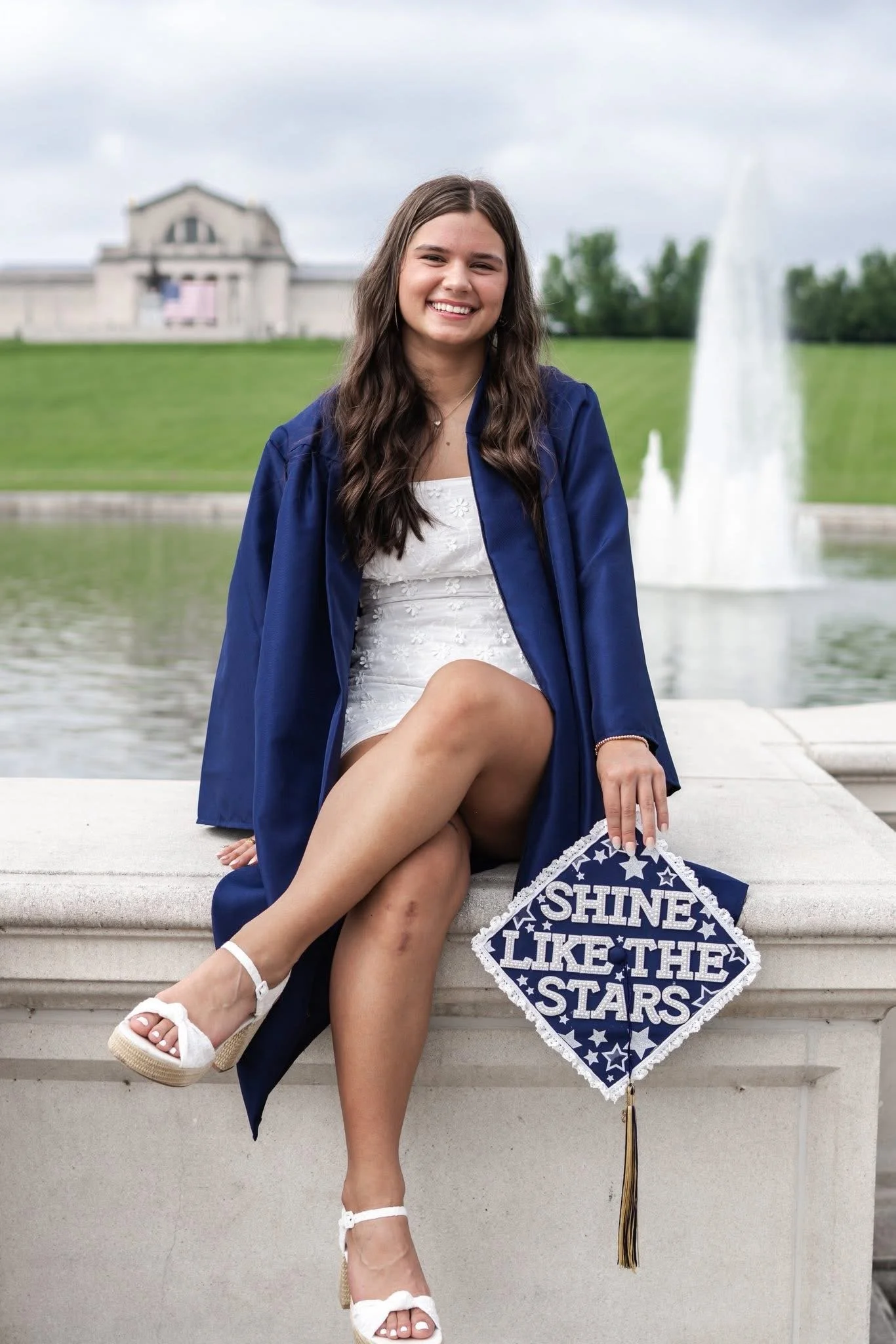 A young woman in a blue graduation gown and sandals sitting on a concrete ledge in front of a fountain, smiling at the camera, holding a sign that says 'Shine Like the Stars' with a tassel hanging from it.
