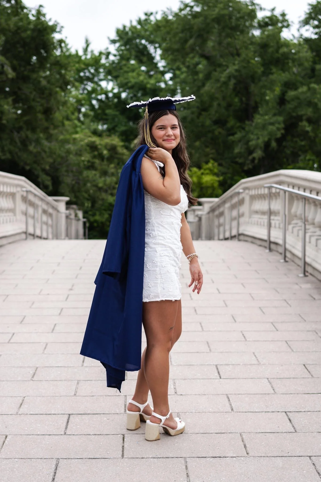 A young woman in a white dress and white platform heels is holding a blue graduation gown over her shoulder. She is standing on a stone bridge, smiling, with a graduation cap on her head and green trees in the background.