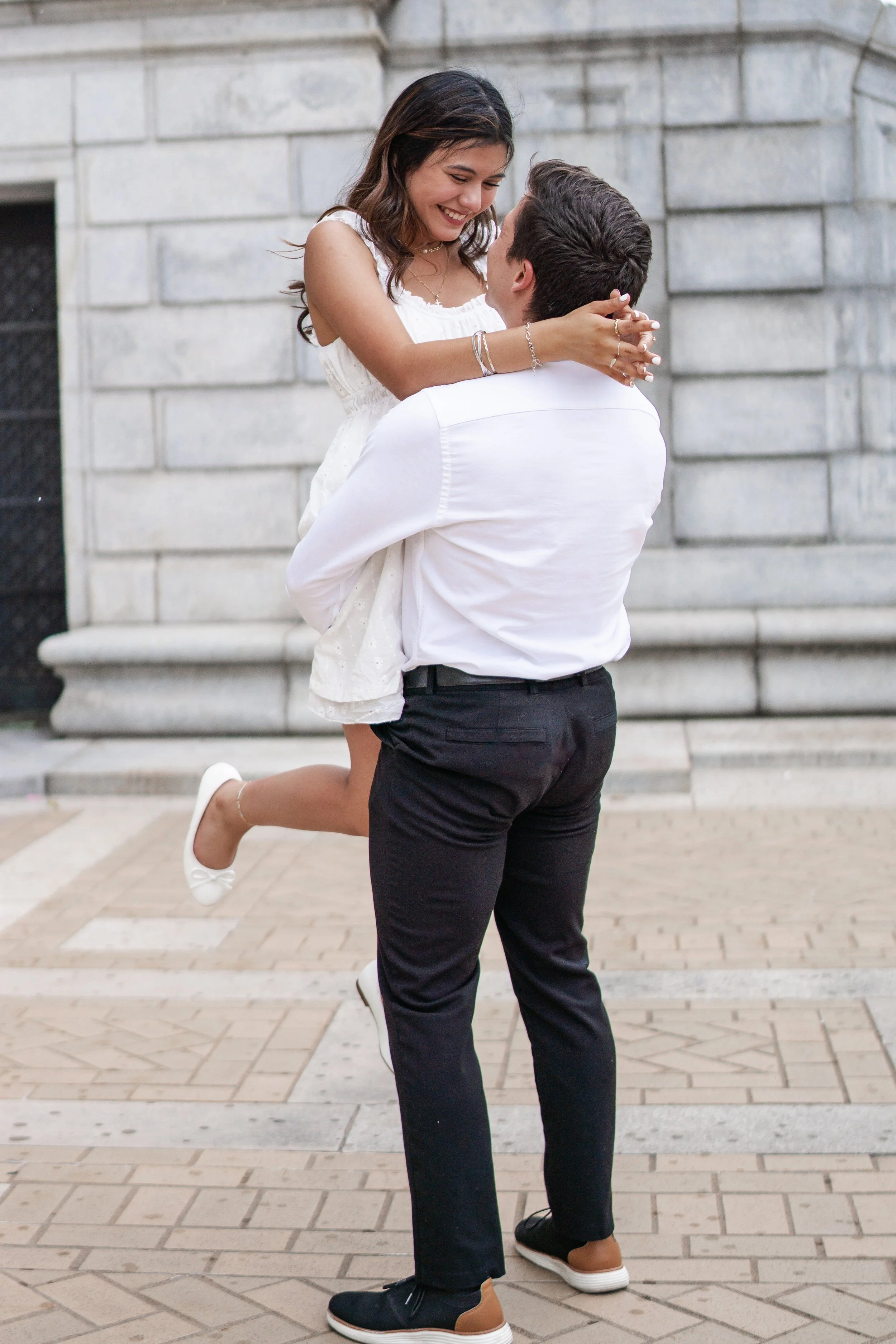 A man in black shoes and black pants is holding a woman in white dress with white shoes, as she jumps, in front of a stone building with steps.