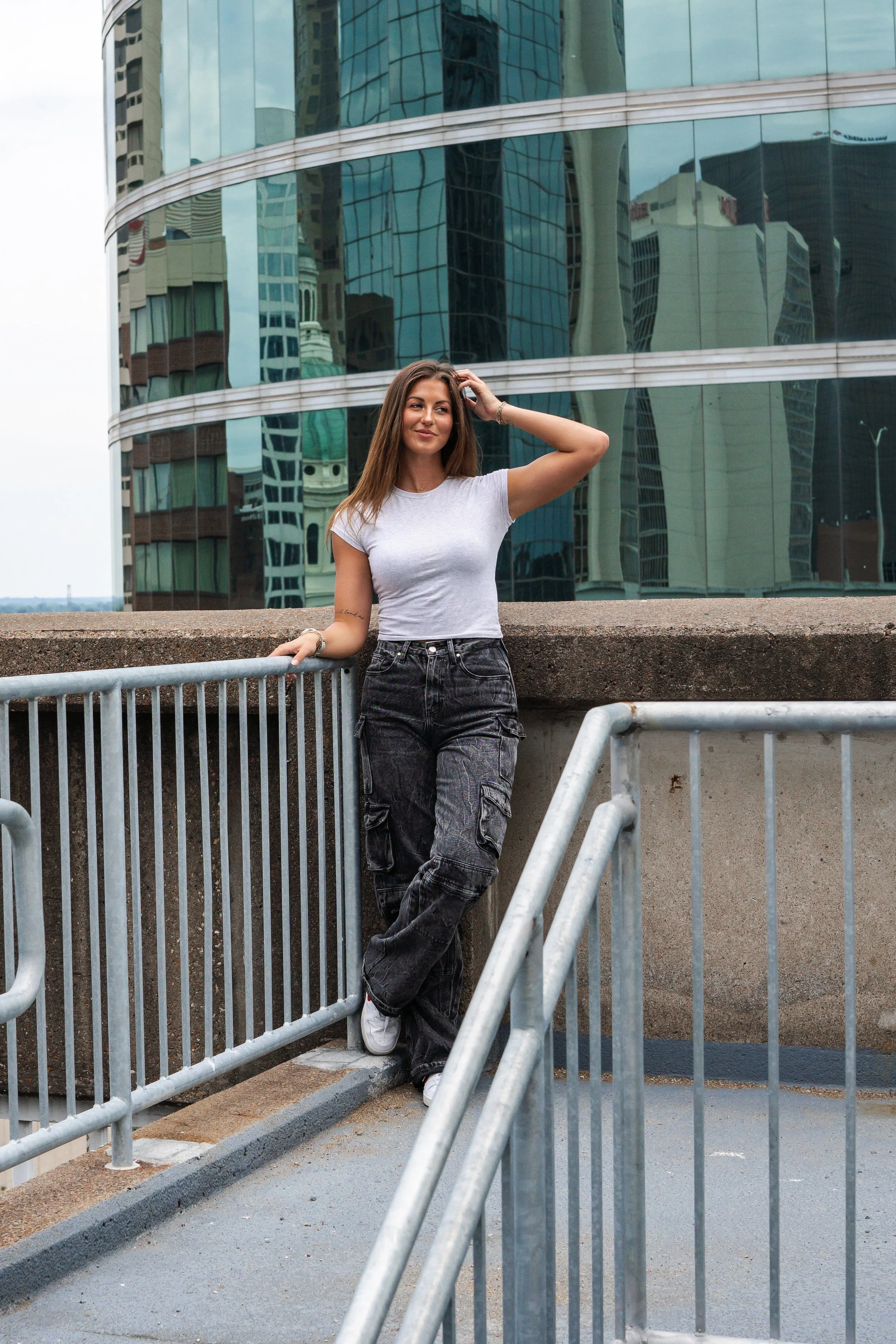Young woman with long brown hair wearing a white t-shirt and cargo pants leaning on a metal railing in front of a modern glass building in an urban setting.