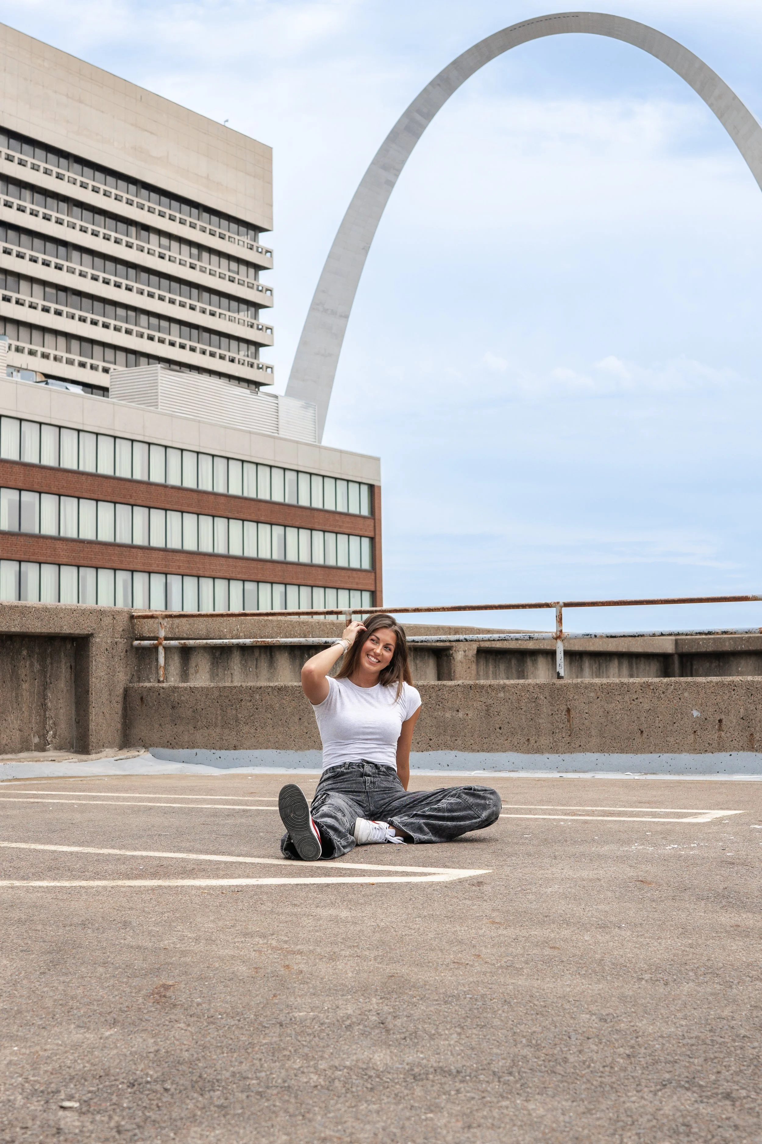 A woman with brown hair and a big smile, wearing a white t-shirt and gray baggy pants, sitting cross-legged on an empty parking lot with the Gateway Arch and a tall building in the background.