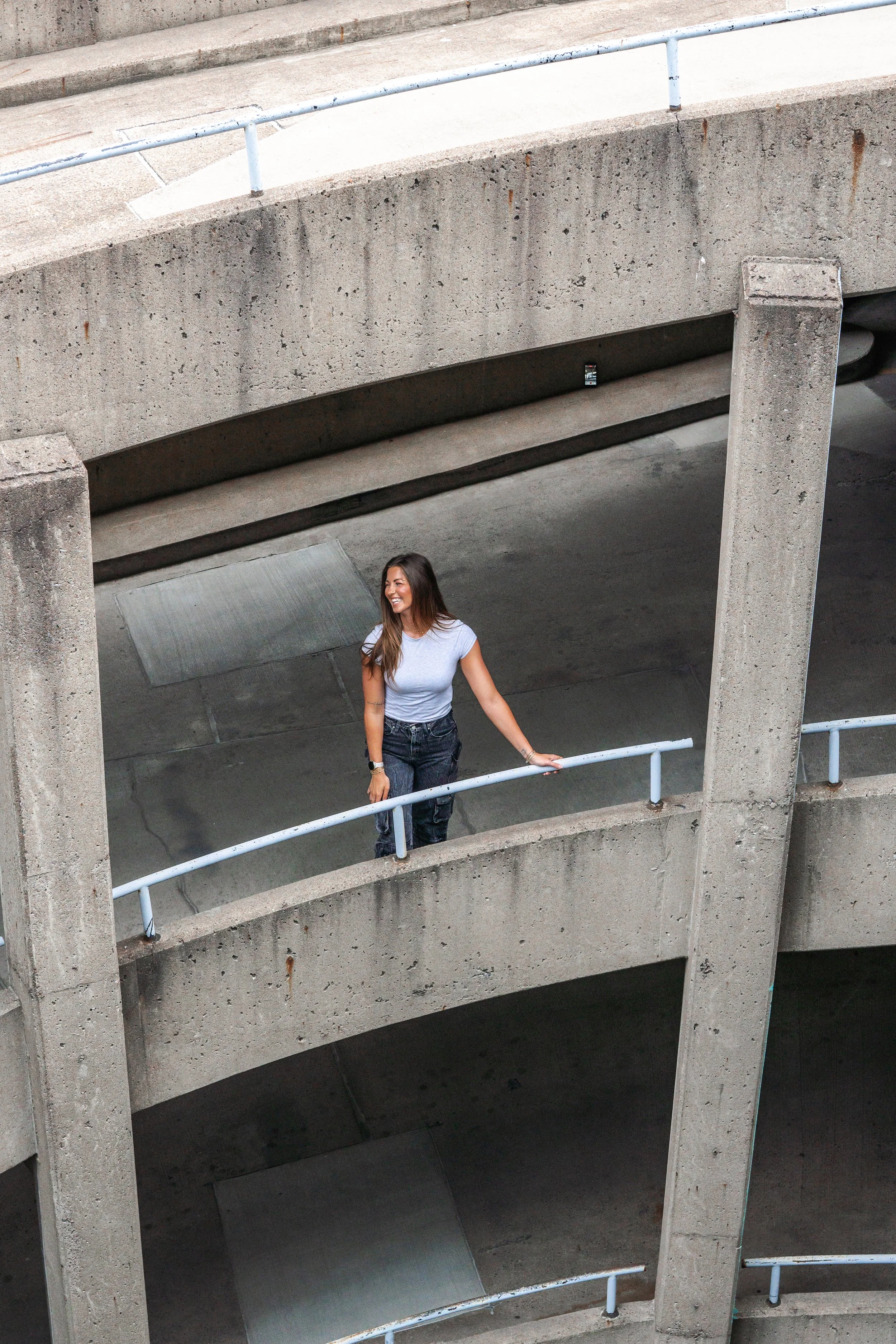 A woman with long brown hair, wearing a light gray t-shirt and black pants, smiling and holding onto a metal railing on a concrete staircase with multiple levels and metal railings.