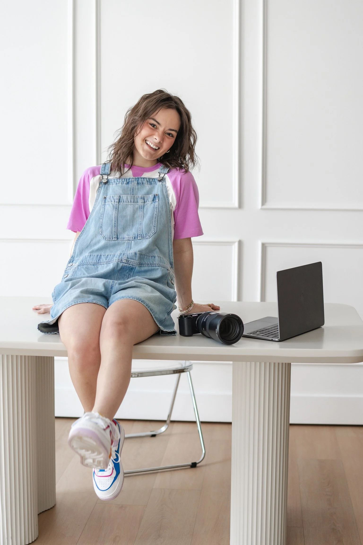 A young woman sitting on a white table with a white background, smiling, wearing a pink t-shirt, denim overalls, white sneakers with colorful accents, with a black camera and a silver laptop on the table.