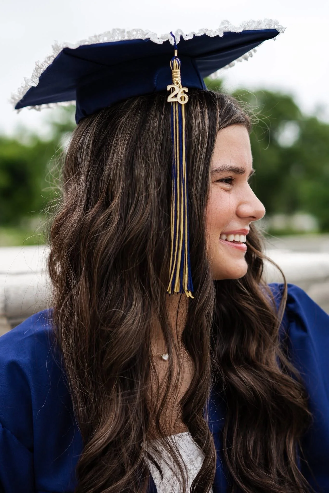 A young woman wearing a blue graduation cap with a gold tassel and a '26' charm, smiling with long brown hair and a white shirt under a blue gown, outdoors with blurred greenery in the background.