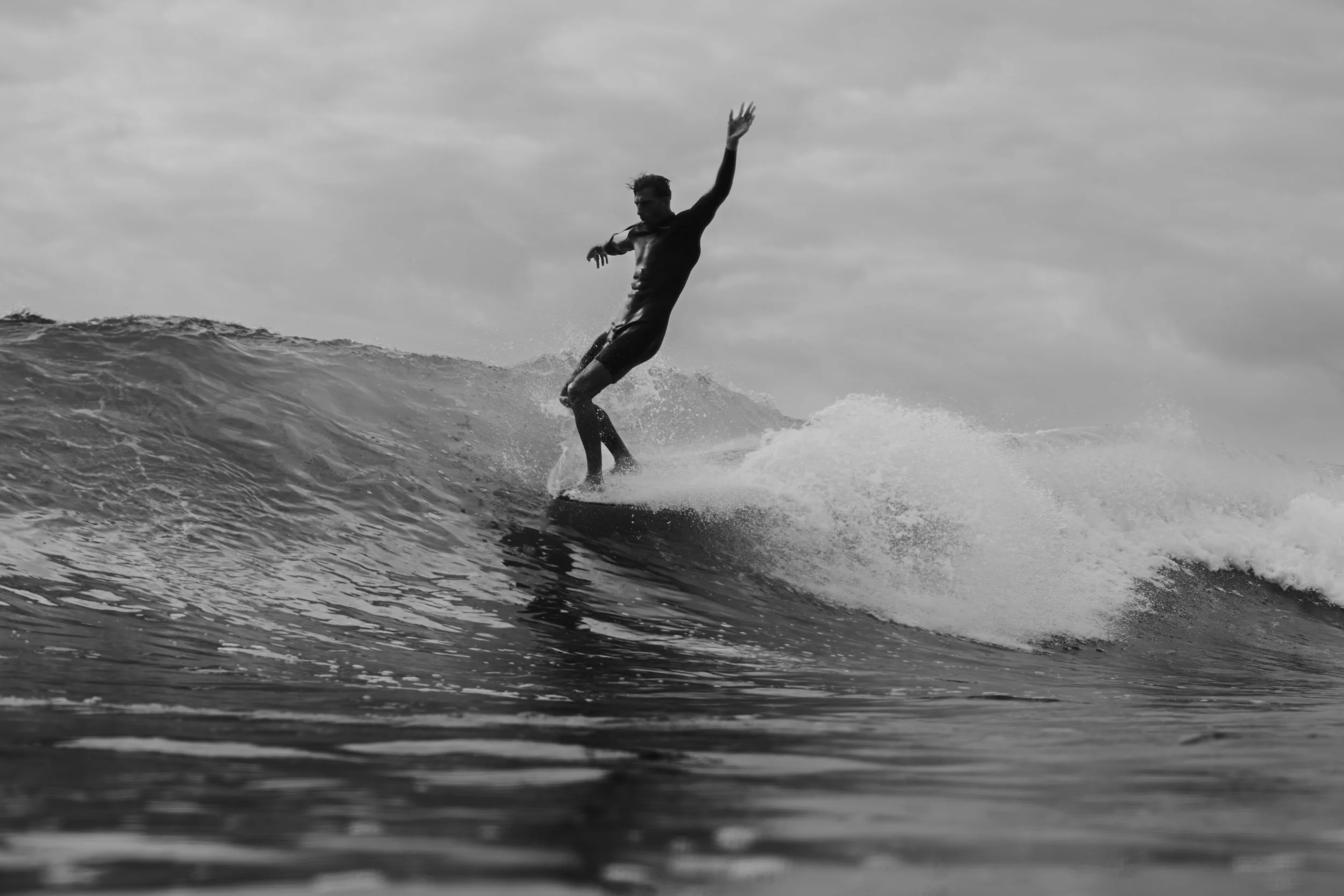 Black and white photo of a man surfing on a wave, with an overcast sky in the background.