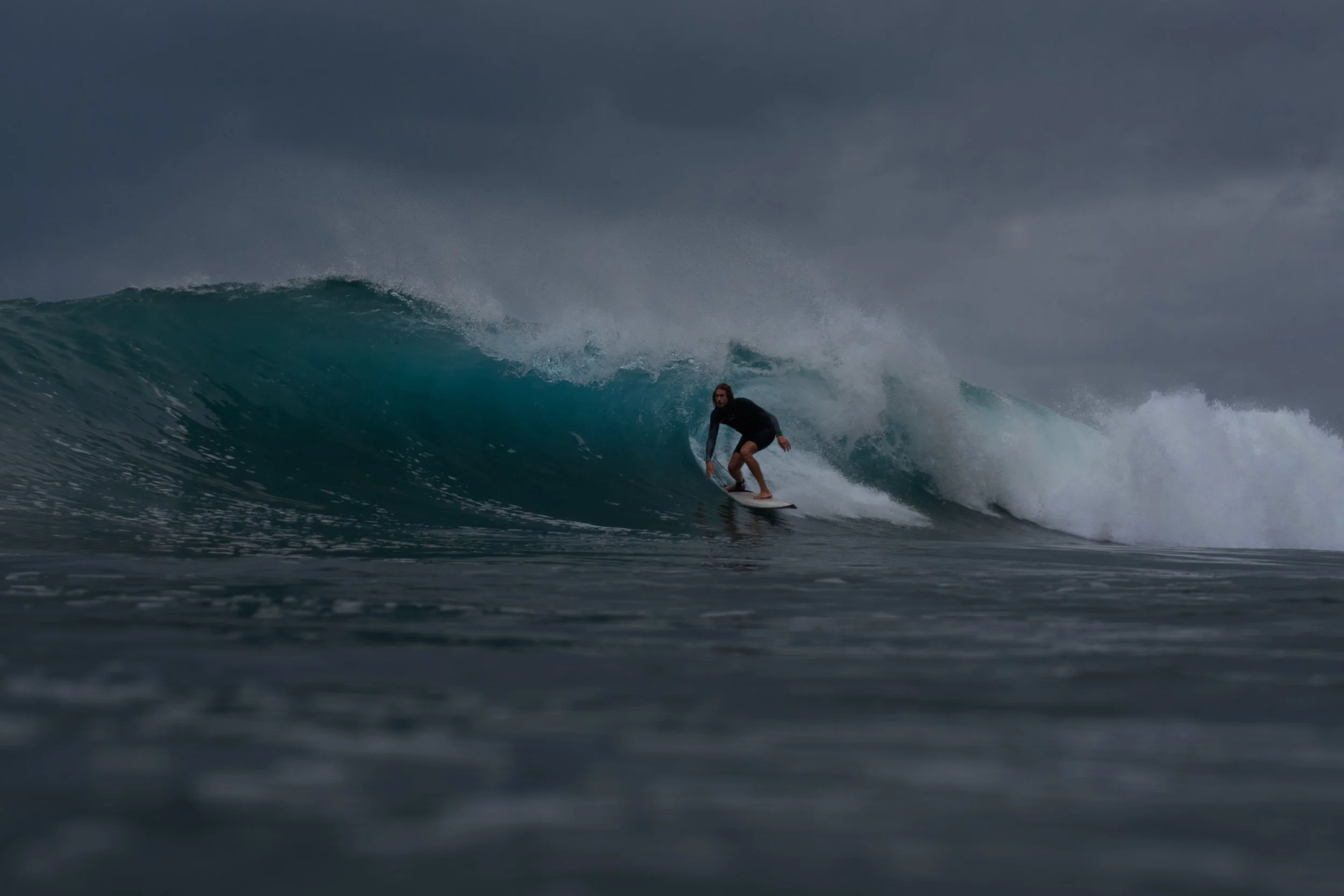 A person surfing on a wave under dark, cloudy skies.