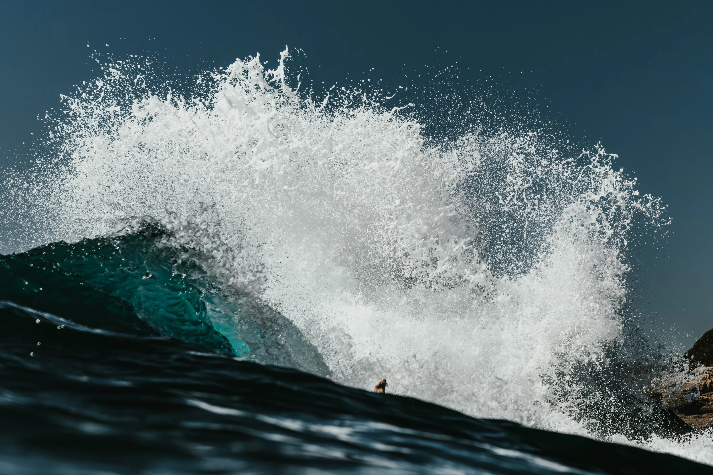 Ocean wave crashing with white spray against a clear blue sky.