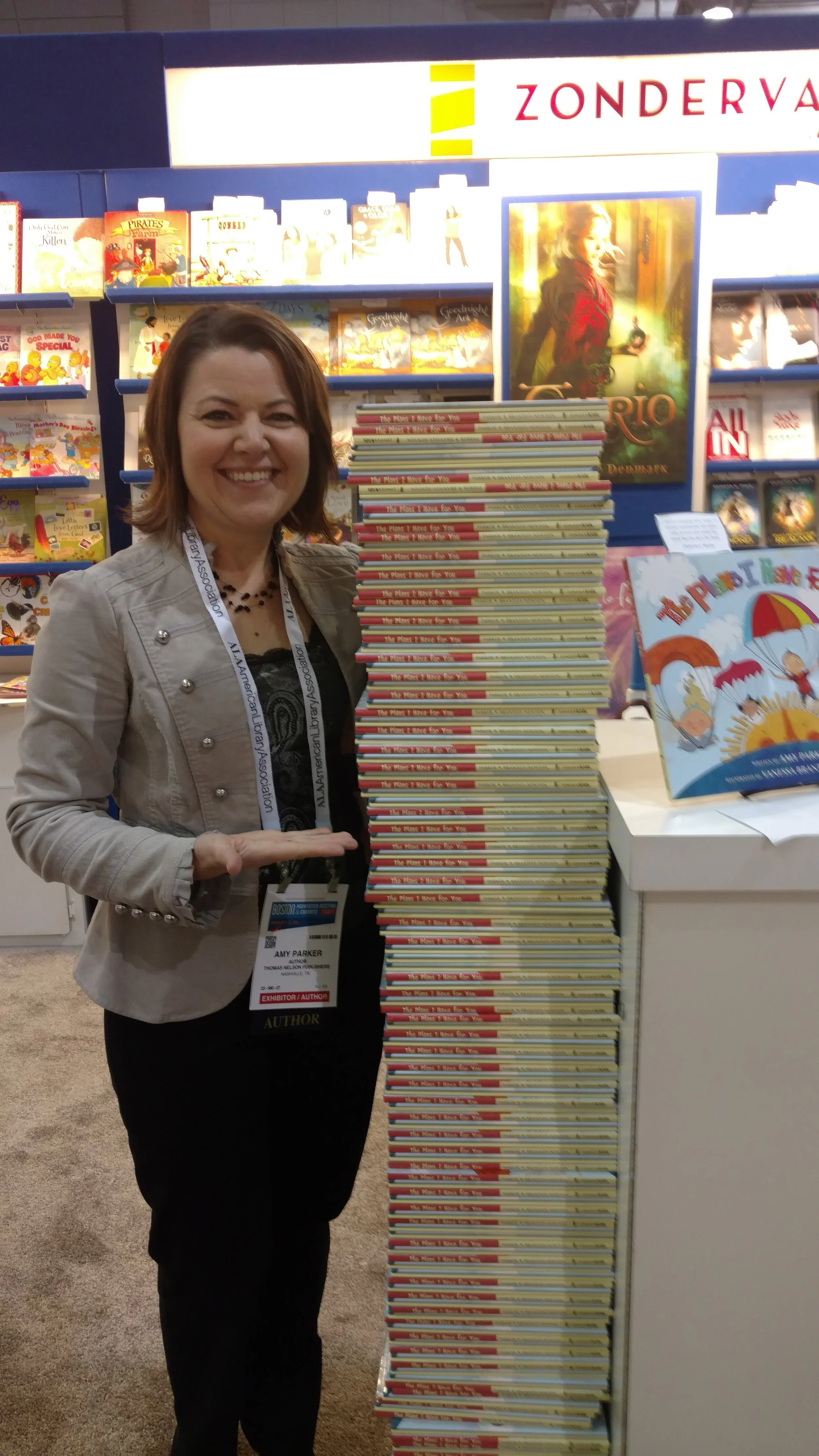 Amy Parker at a book fair stands next to a tall stack of books titled "The Plums I Have For You." The background includes bookshelves with various children's books.