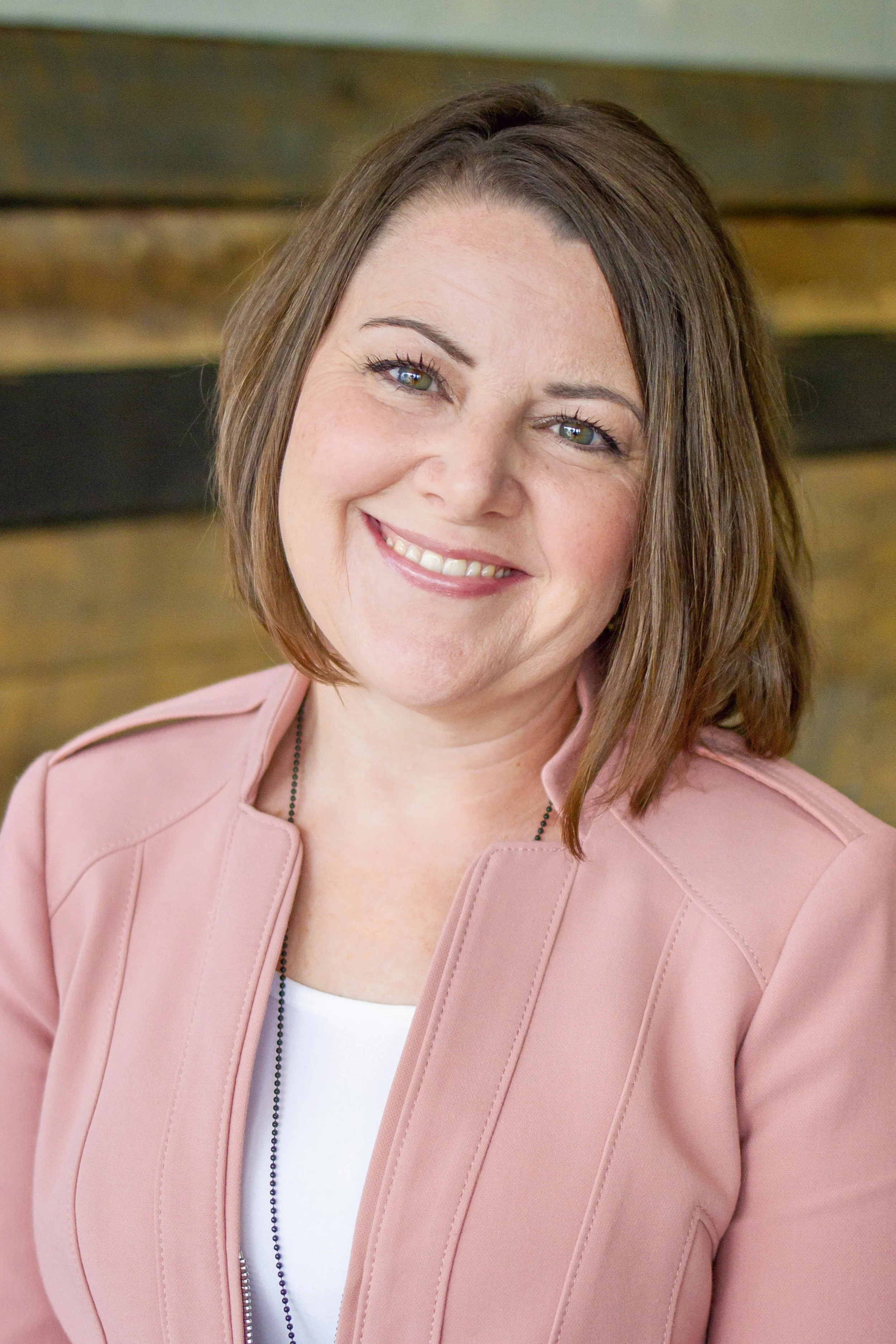 A woman with shoulder-length brown hair wearing a pink jacket and a white top, smiling, with a blurred background.