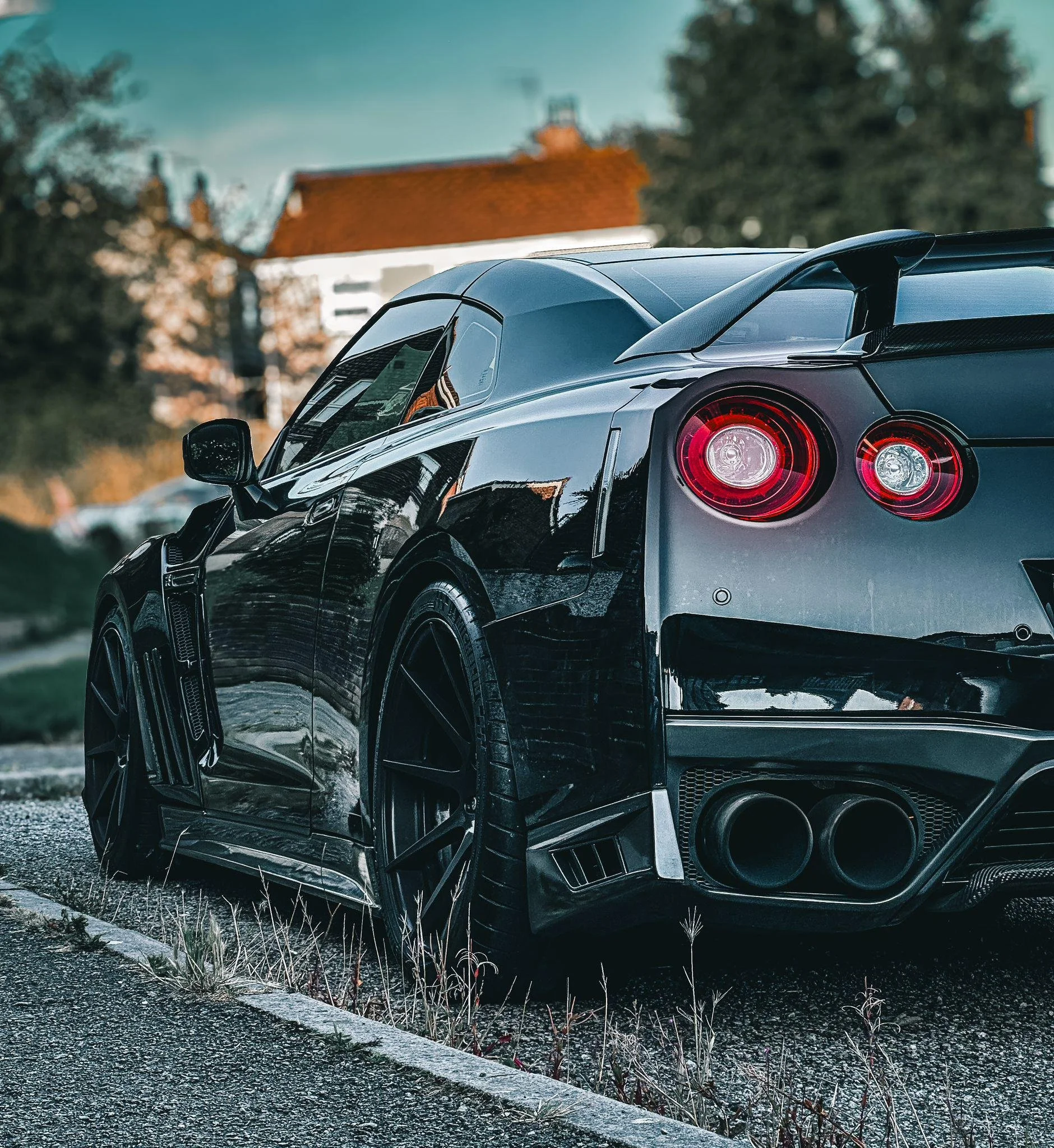 Close-up of a silver sports car with black accents, parked on a gravel surface near some weeds, with trees and a building with a red roof in the background.