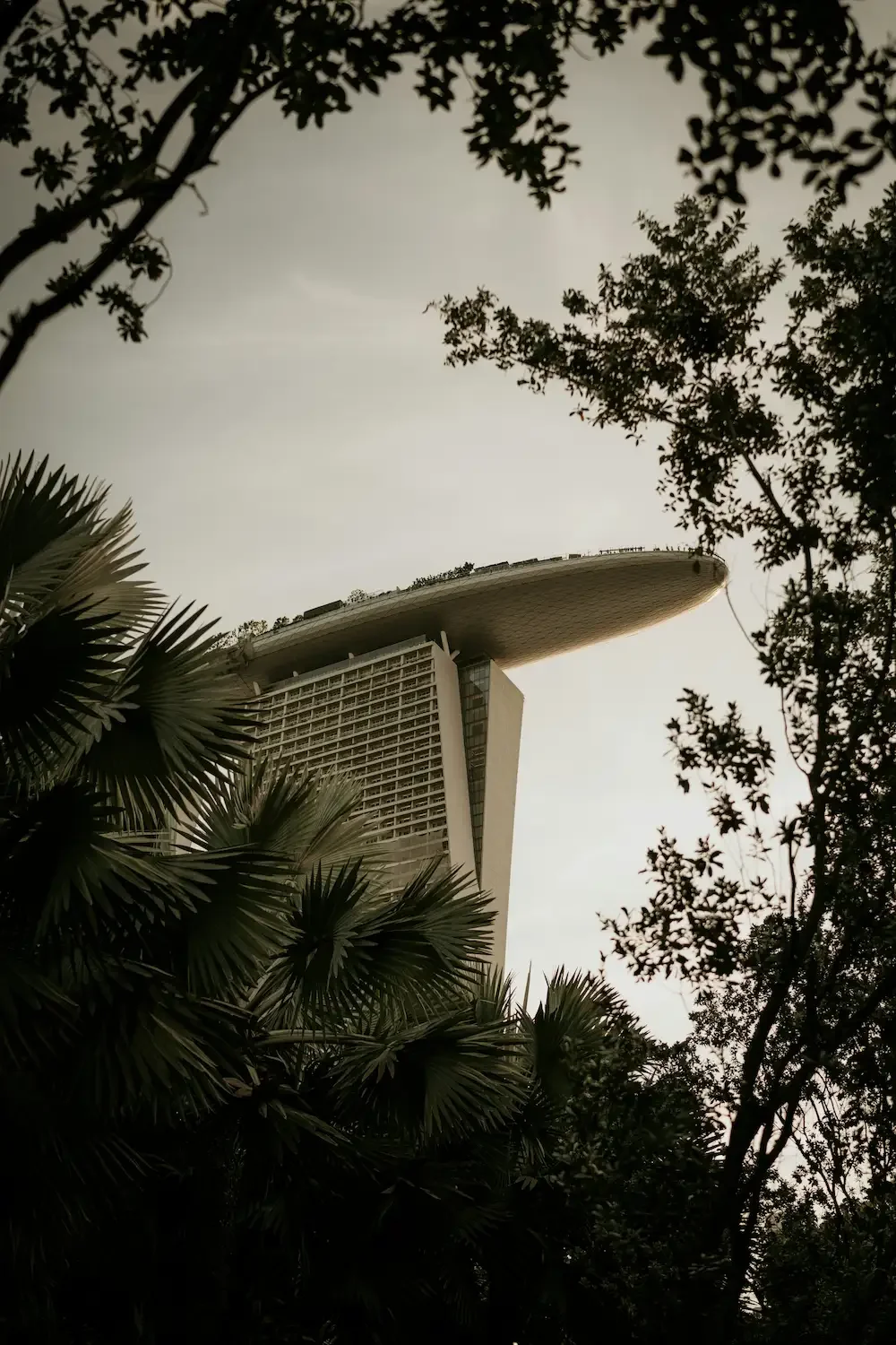 A low-angle view of the Marina Bay Sands hotel in Singapore, with the iconic three-towered structure topped by a boat-shaped sky park, partially obscured by surrounding trees and foliage.