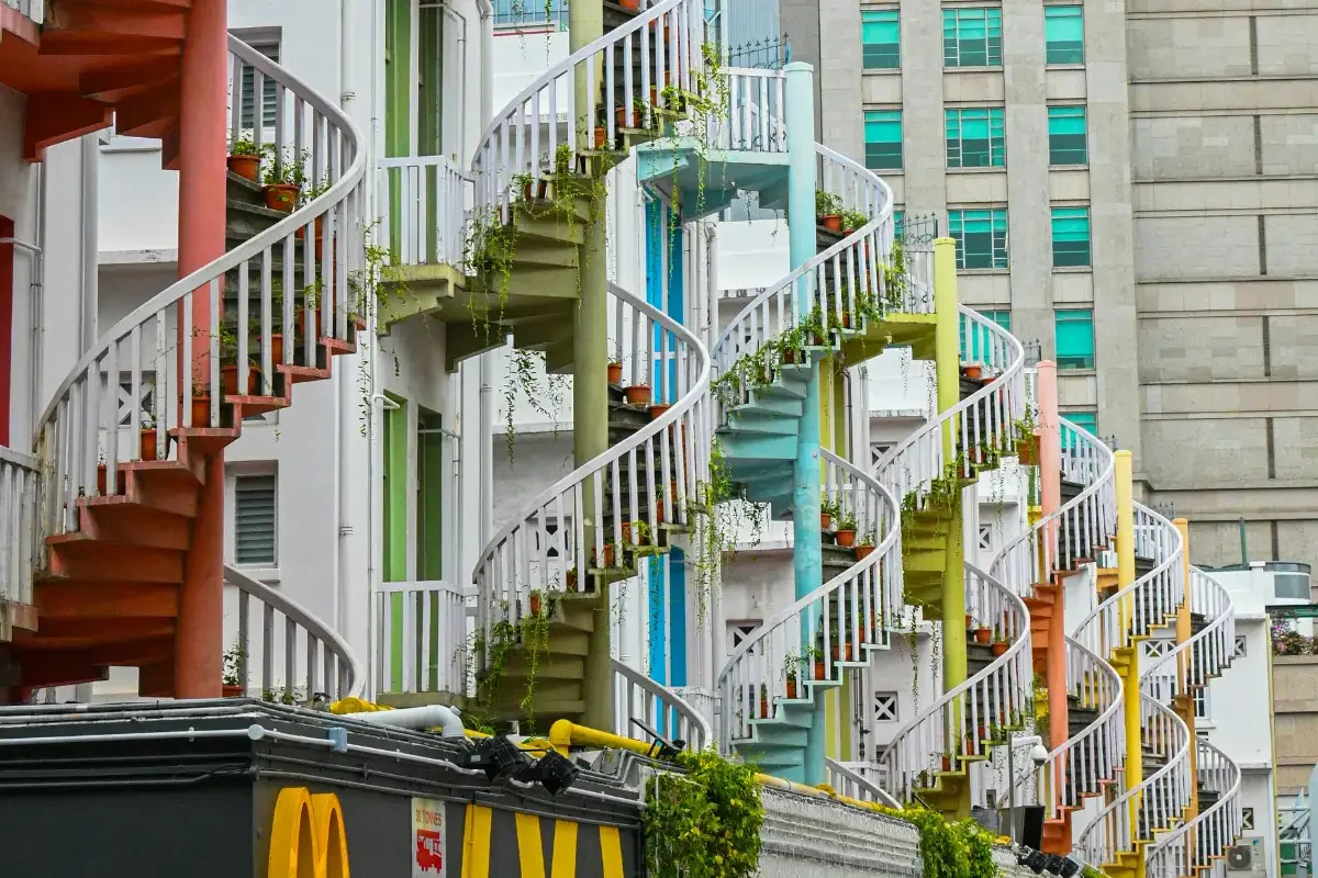Colorful outdoor spiral staircase with potted plants on each level, set against a building with blue windows.