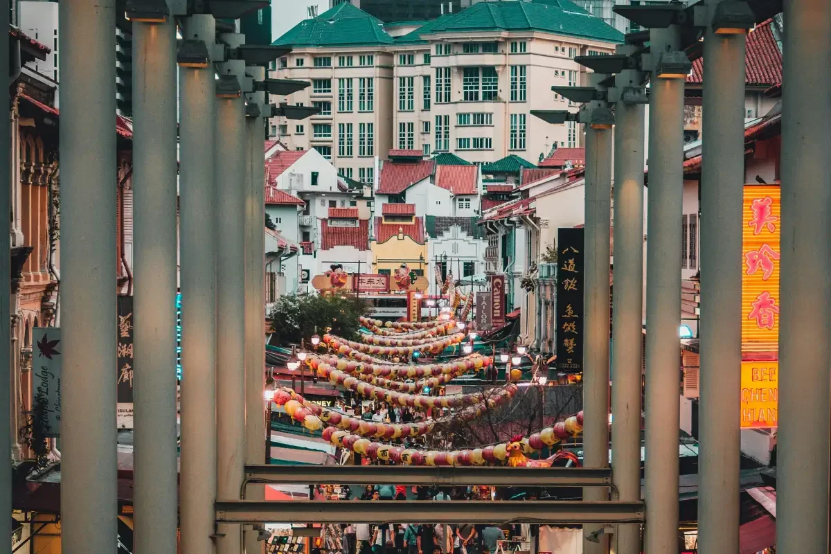 View of a bustling street decorated with colorful lanterns and signs, framed by vertical metal bars, with buildings and multi-level structures in the background.