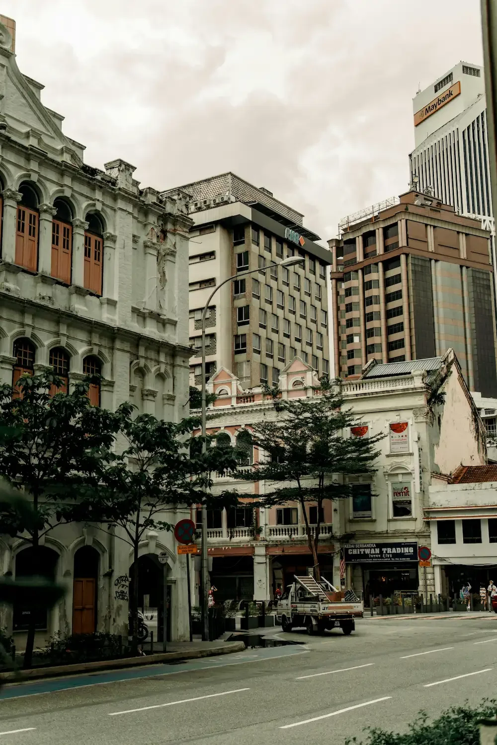 City street scene with old white colonial-style buildings, modern skyscrapers in the background, trees, and a parked utility vehicle.