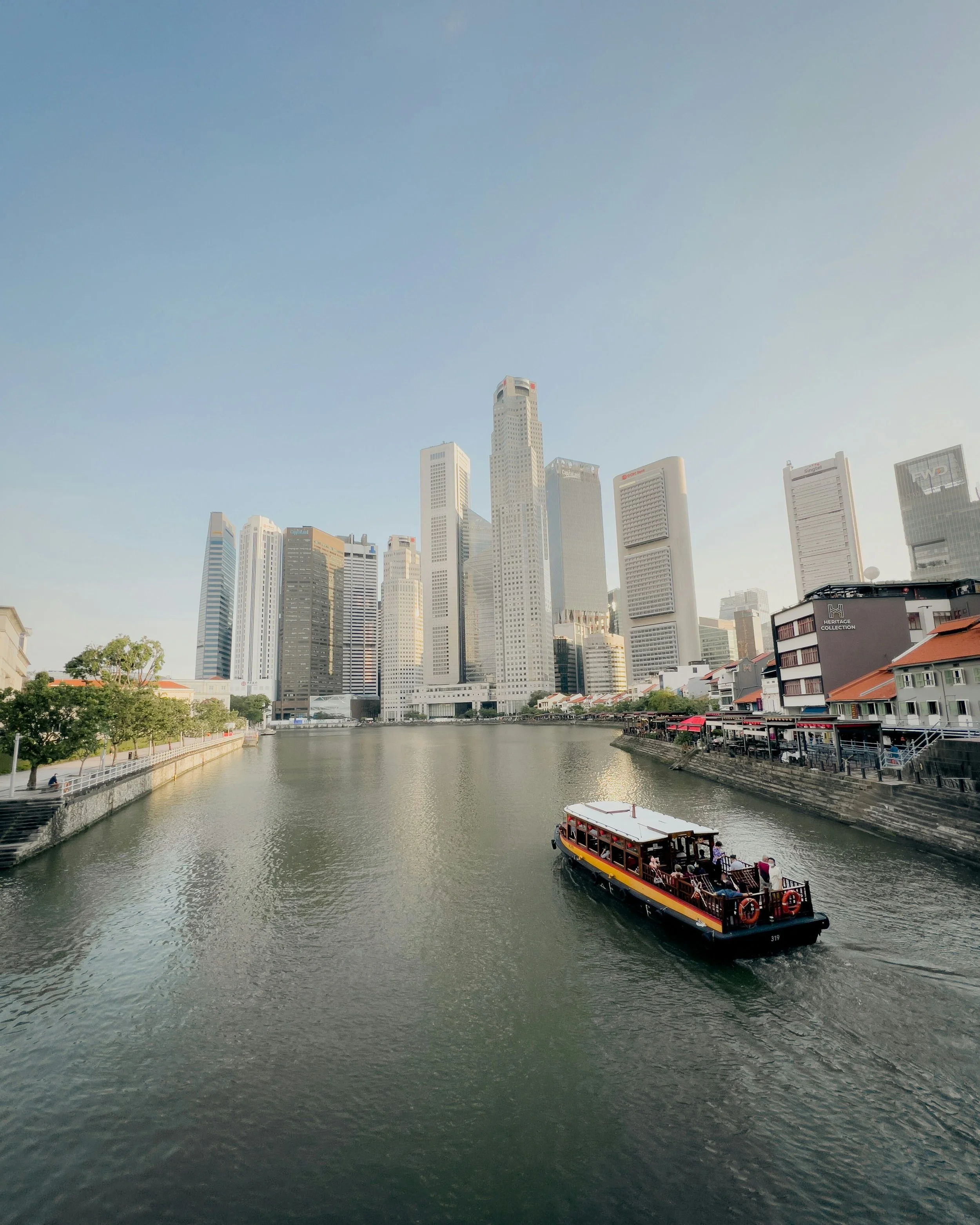City skyline with tall skyscrapers and a river in the foreground. A boat with tourists is sailing on the river.