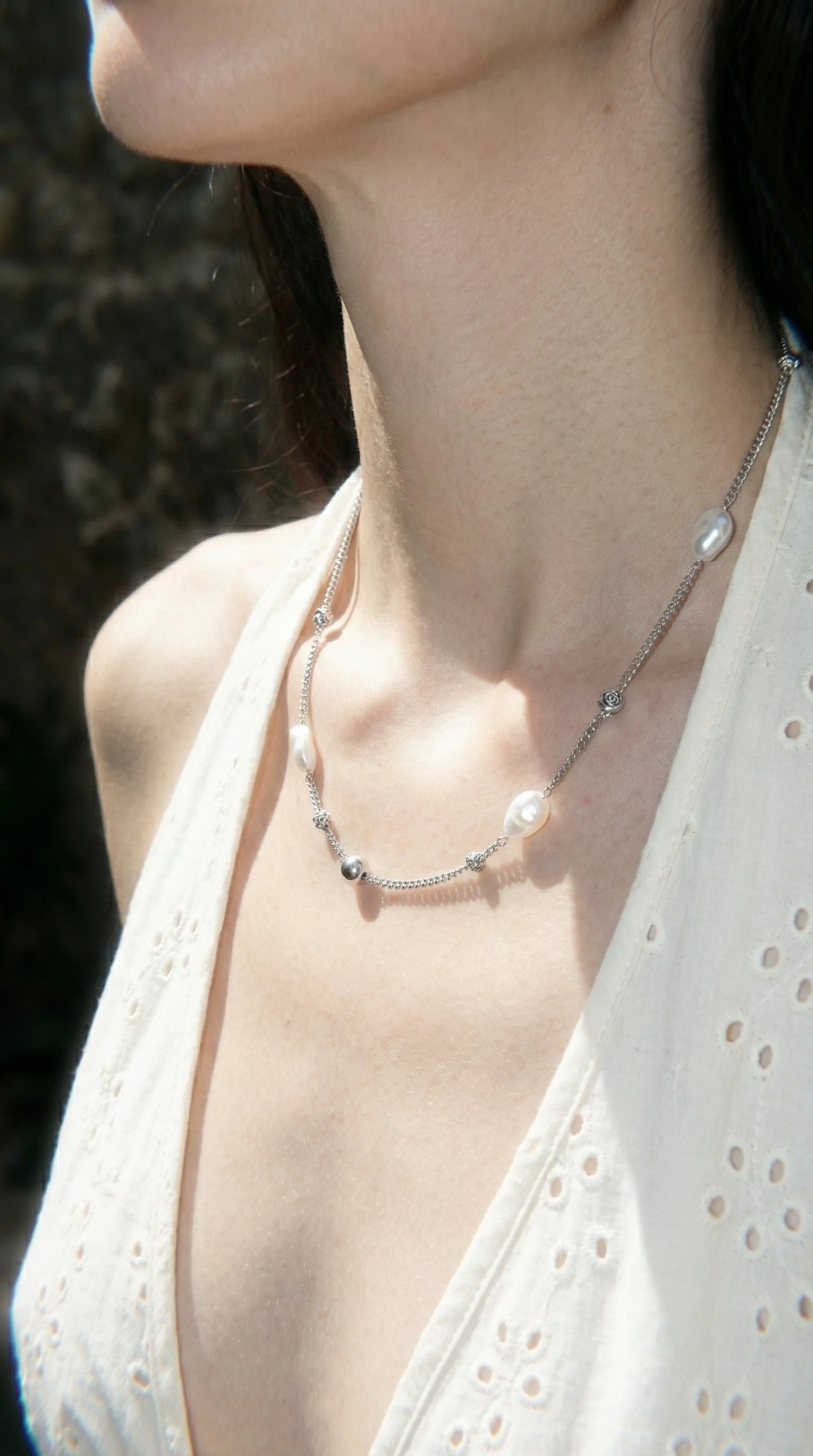 Close-up of a woman's neck and collarbone showing a silver necklace with pearl and gemstone embellishments, wearing a sleeveless cream-colored top with eyelet details.