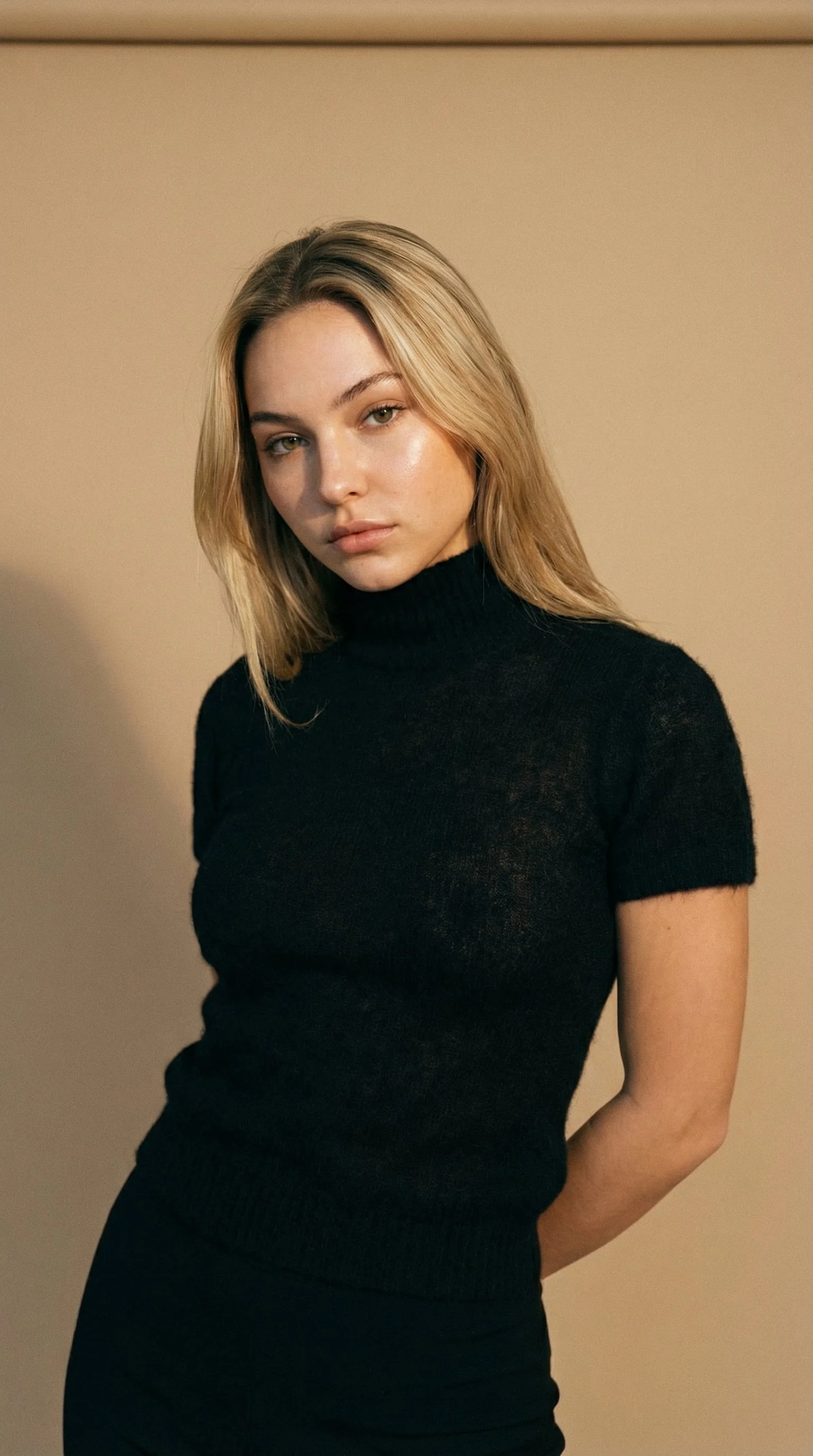 A young woman with blonde hair wearing a black turtleneck sweater, standing against a beige wall.