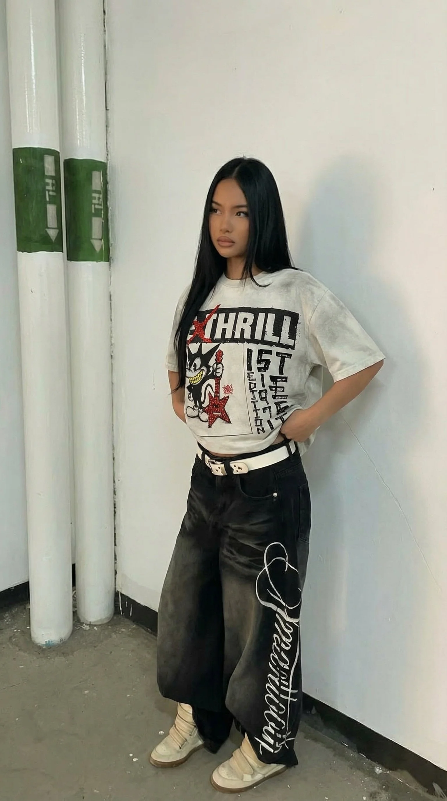 A woman with long black hair wearing a graphic t-shirt and baggy pants stands against a white wall next to green and white pvc pipes.