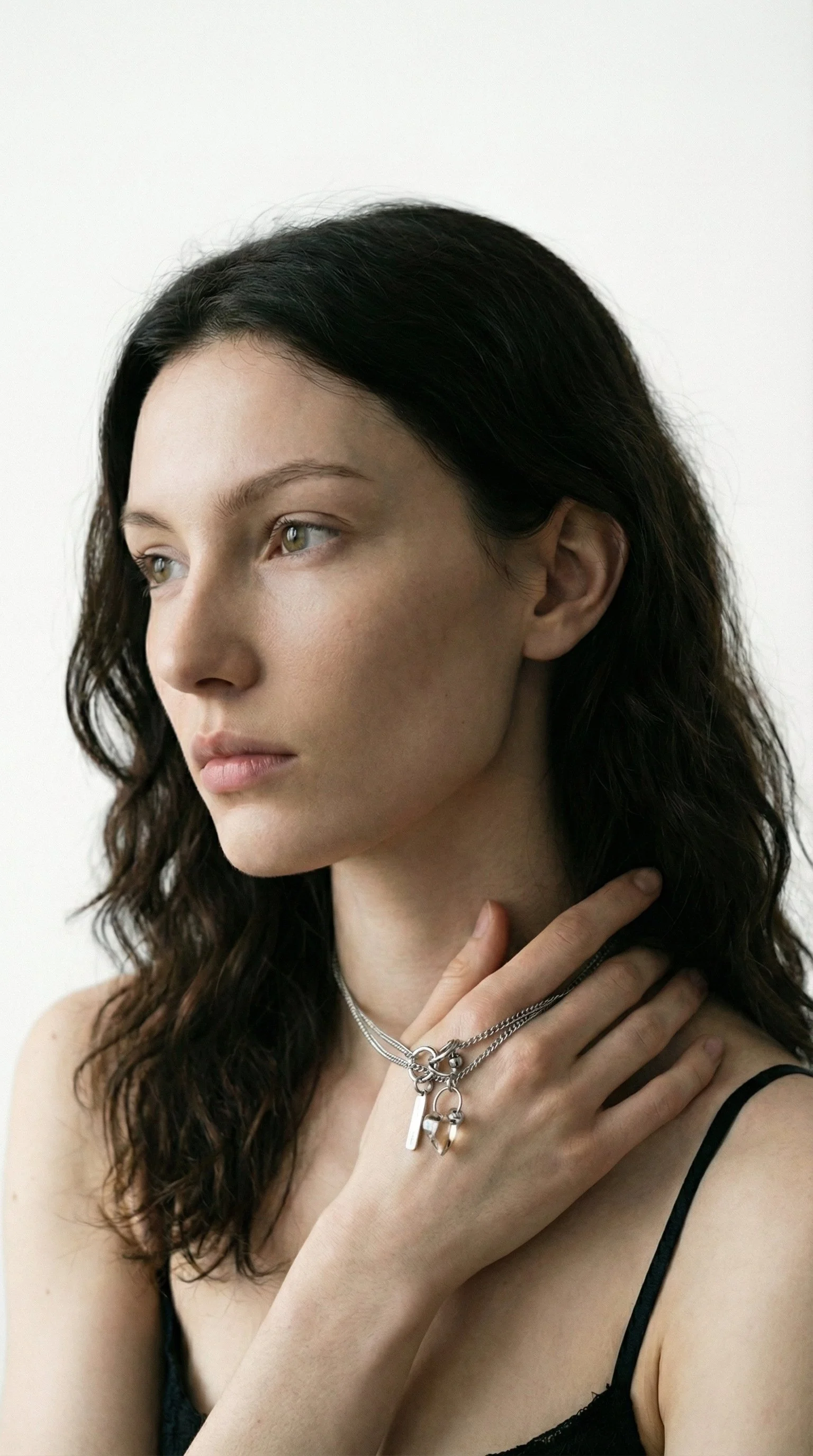 A young woman with wavy black hair looking to the left, wearing layered silver necklaces and a black spaghetti strap top, against a white background.