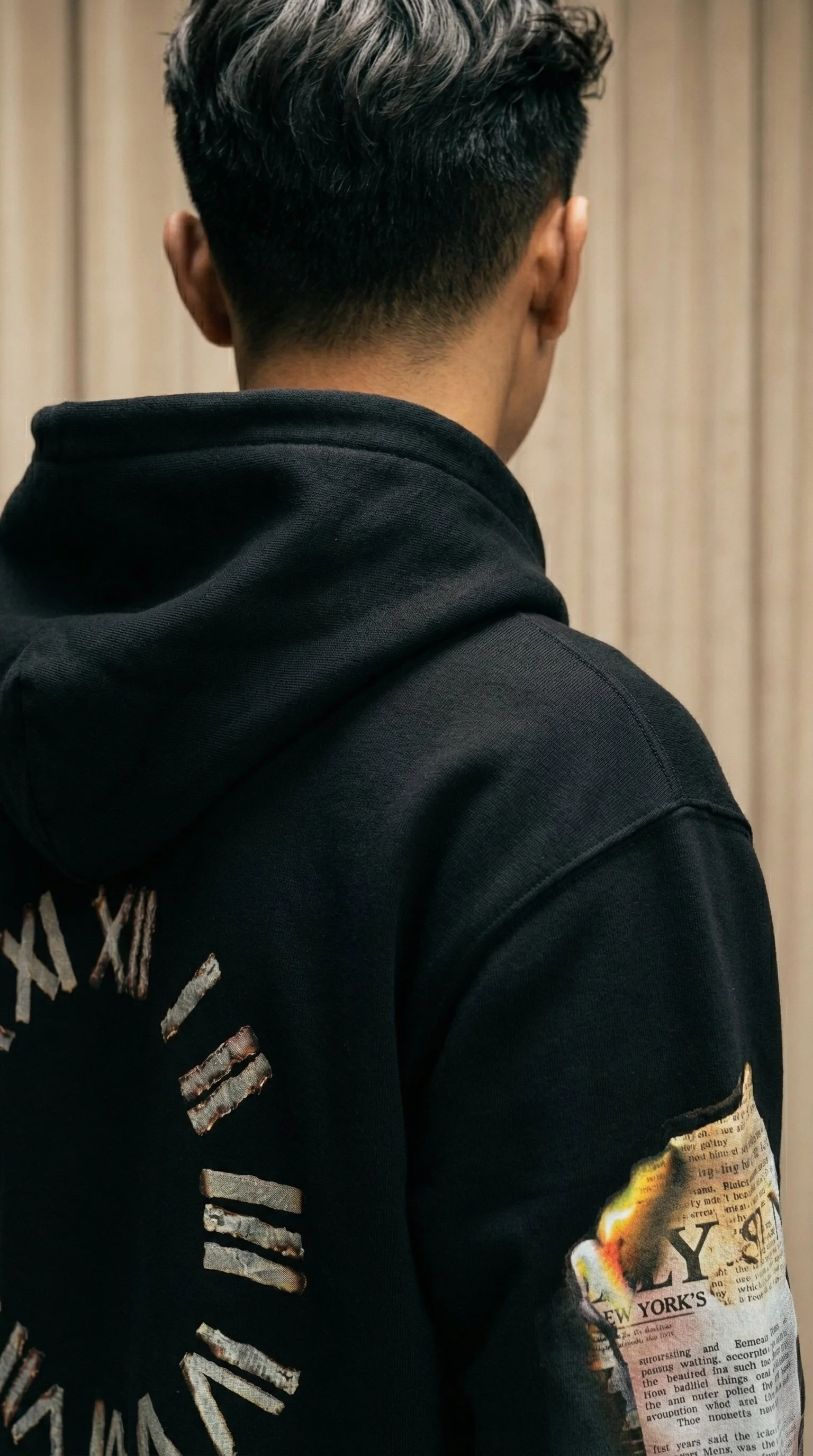 Back of a person wearing a black hoodie with graphic text and newspaper print on the sleeve, standing against a wooden wall.