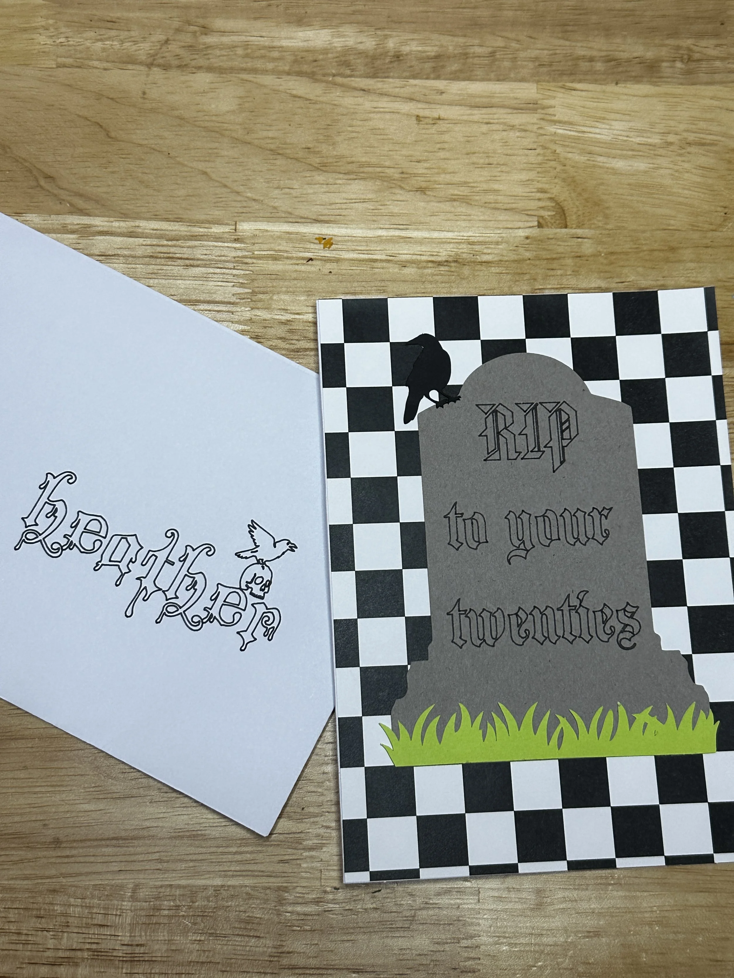 Photograph showing a black and white checkerboard pattern on a card, with a gray tombstone-shaped overlay with words, a black crow silhouette, and some green grass at the bottom. The card also has text that says 'to your' and 'twenties'.
