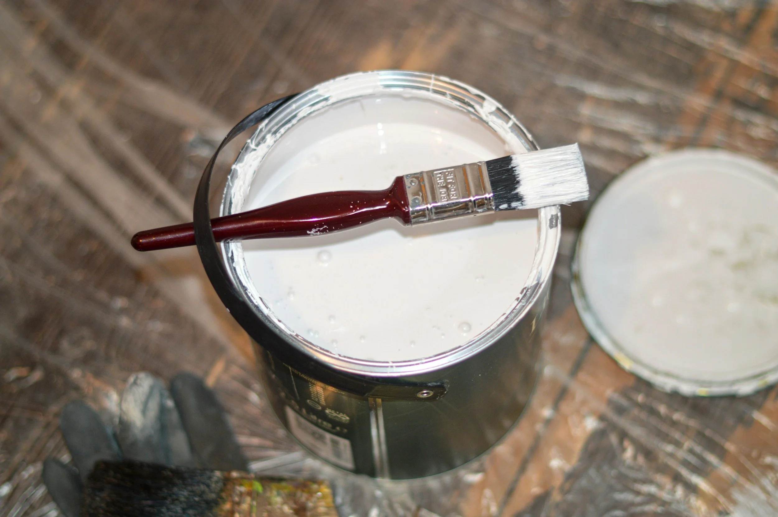 A paintbrush resting on the rim of a container filled with white paint, with another open container of white paint visible in the background. The scene appears to be outdoors on a paved surface.