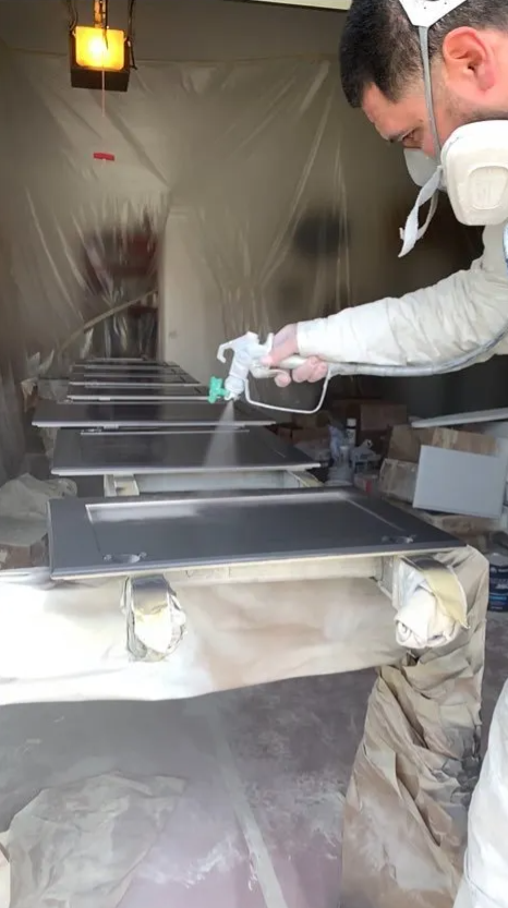A person wearing protective gear, including a mask and gloves, spray-painting or coating a metal surface in a workshop or spray booth.