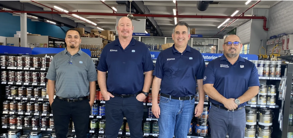 The Kasparian's Paint Center team is shown here in their store. Four men in navy blue uniform shirts smile at the camera in front of a shelf stocked with paint cans.