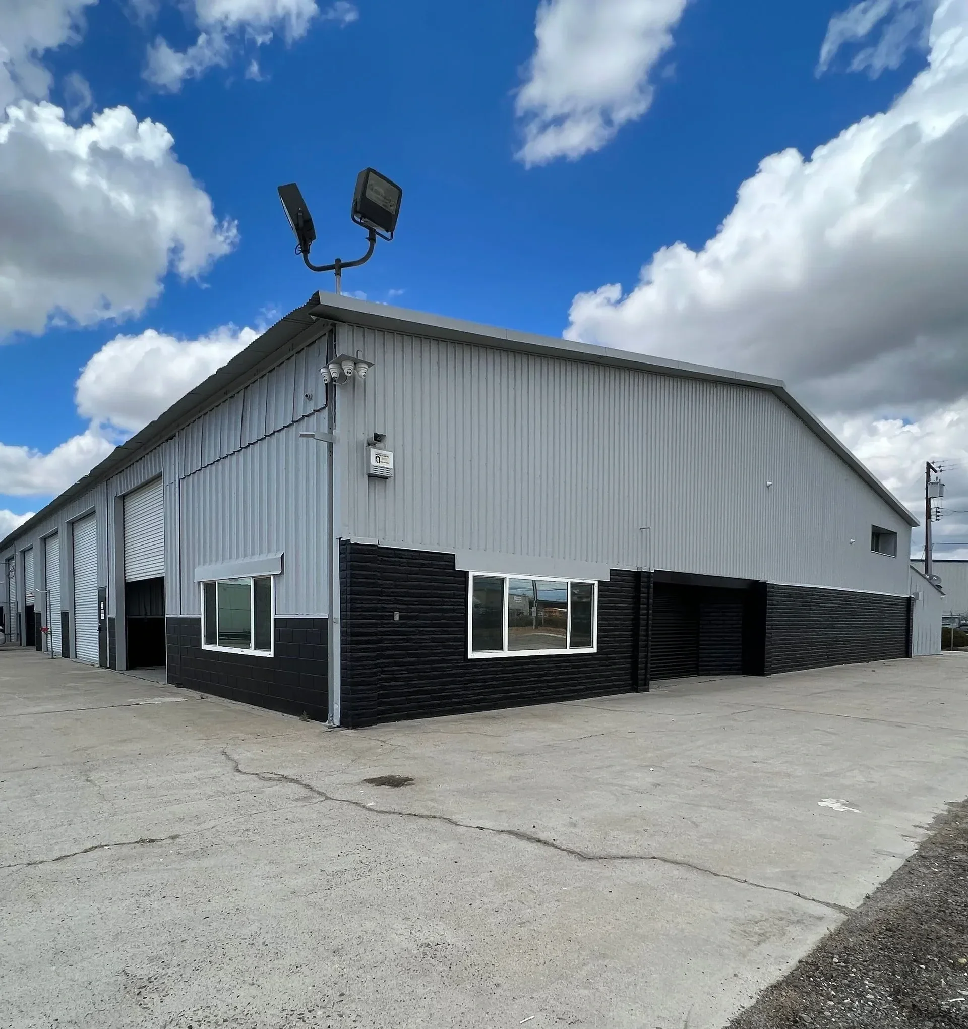 Exterior of a commercial building with metal siding, black accents, and large windows, situated on a paved lot. Security cameras are mounted near the corner of the building, and floodlights are installed on the roof. The sky is partly cloudy with patches of blue visible.