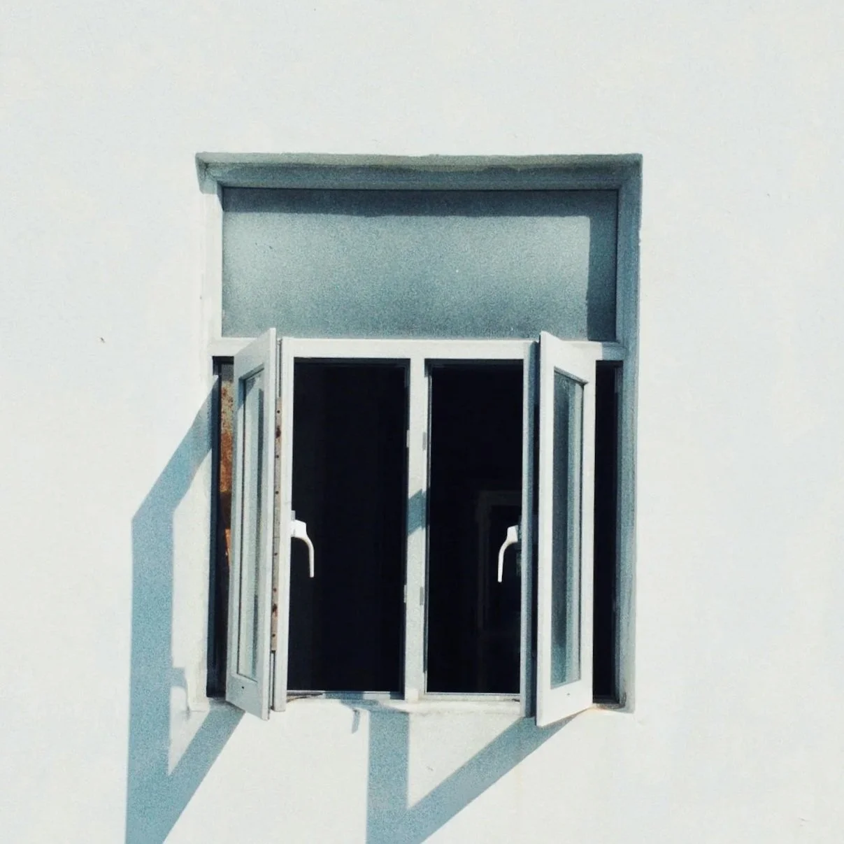A small window with open white shutters on a white wall, casting shadows.