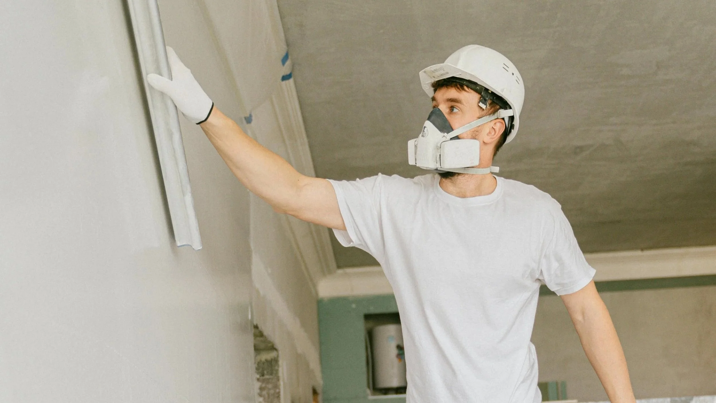 A man wearing a white safety helmet, face mask, white T-shirt, and gloves is painting a wall in a room, using a brush.