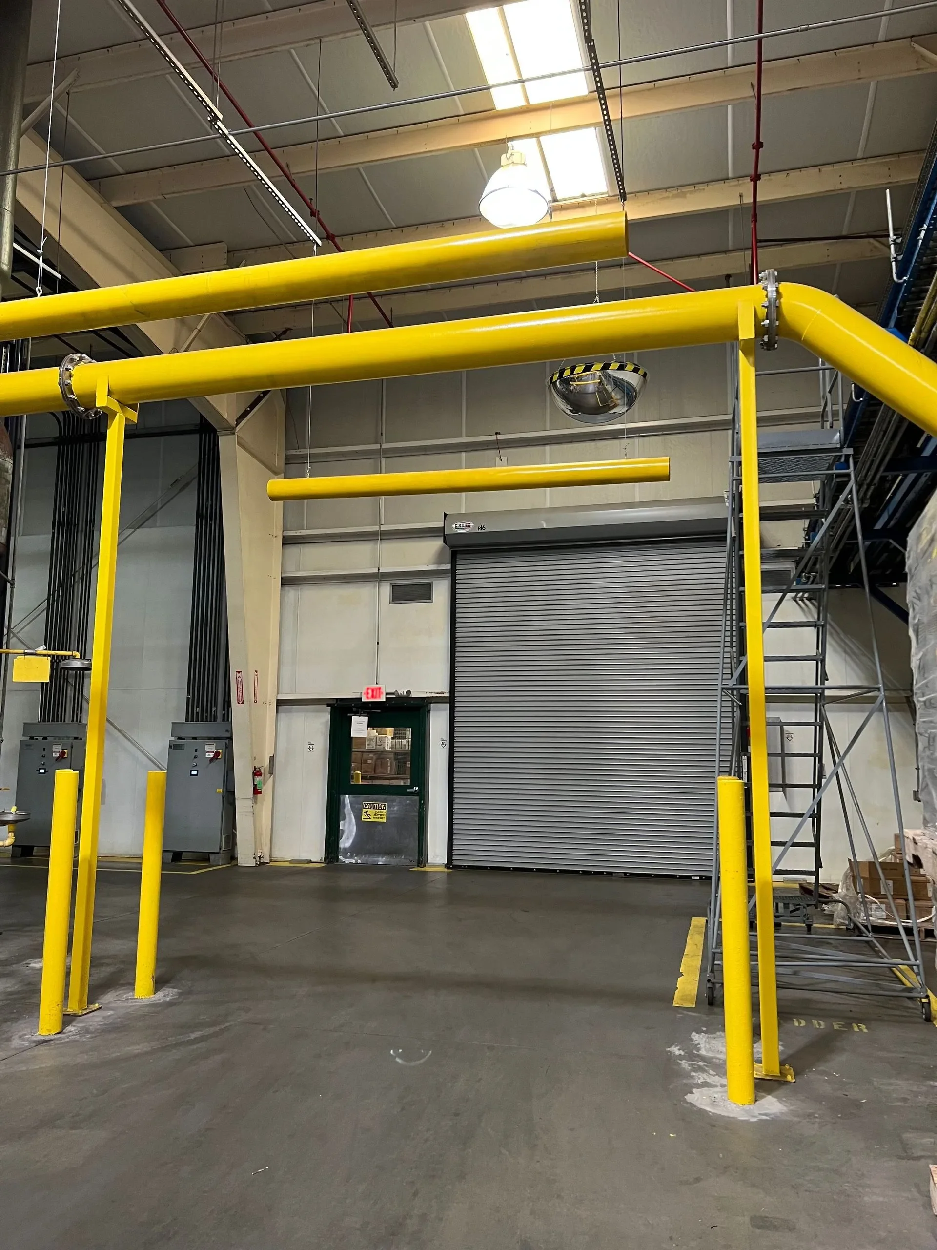 Industrial interior with yellow safety pipes, a metal staircase, a rolling door, and electrical boxes.