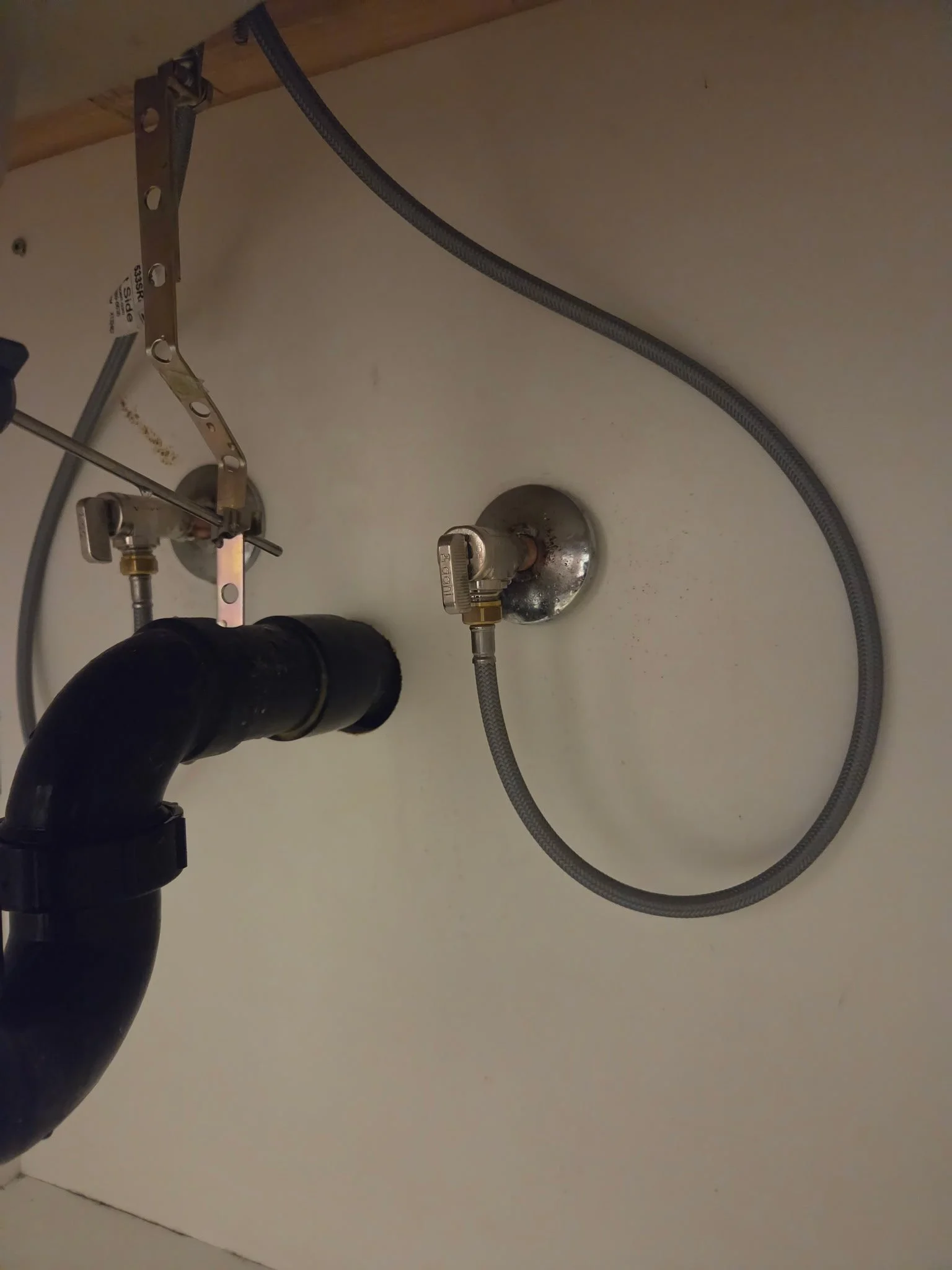 Underneath a kitchen sink, there is a black pipe connected to the drain, a flexible metal supply line, and a metal shut-off valve attached to a wall.