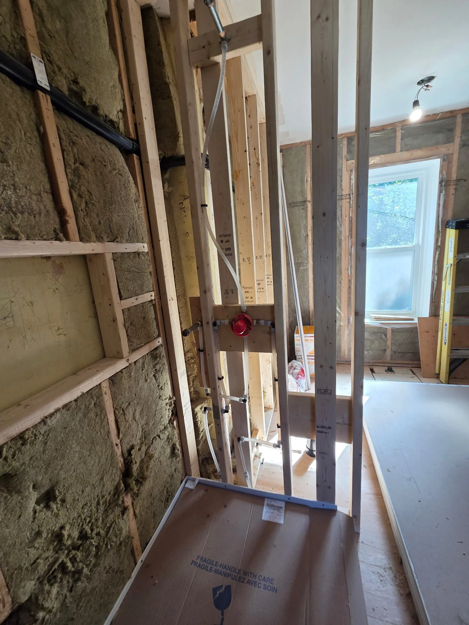 Construction site showing exposed wall studs, piping, insulation, and a window, with a ladder and a partially installed ceiling.