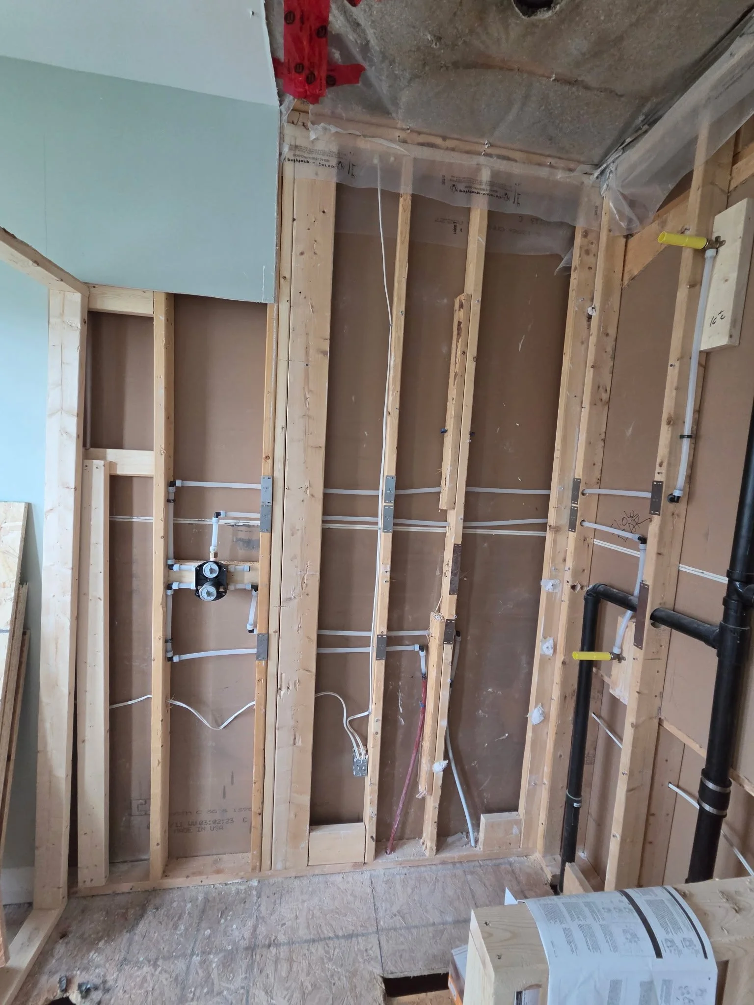 Interior view of a construction site showing wooden framing for walls, electrical wiring, and plumbing pipes. Part of the wall is covered with green drywall, with tools and construction materials visible.