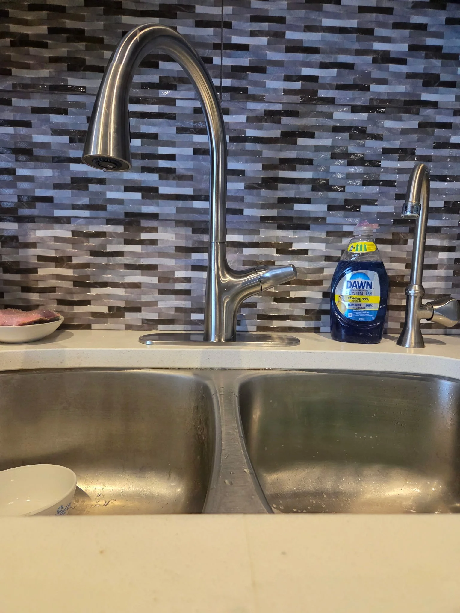 Double kitchen sink with a stainless steel faucet, a dish soap bottle, a bowl with some food, and a cup, against a gray and black tiled backsplash.