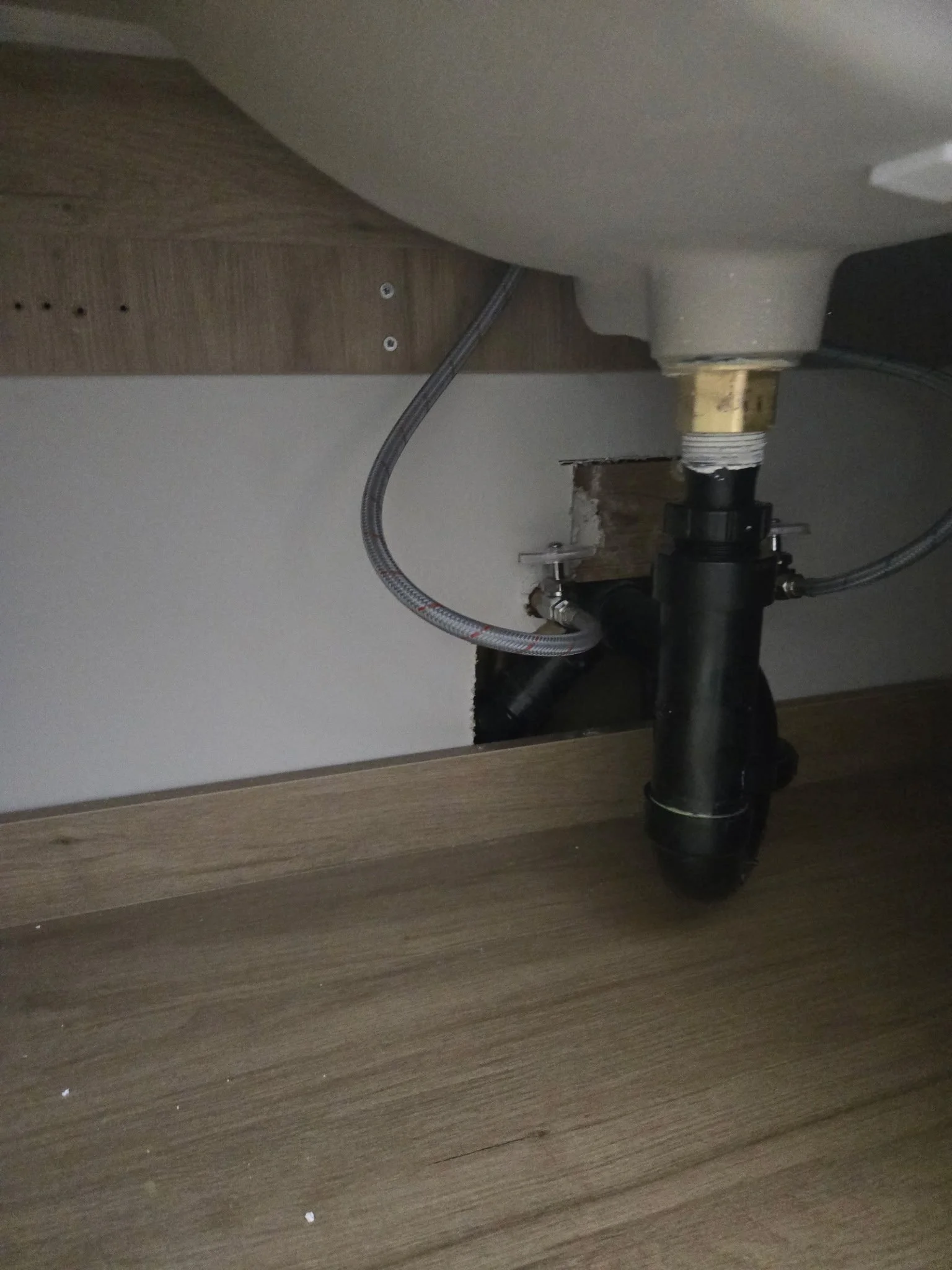 Under a kitchen sink, pipes and hoses are visible, with a section of the wooden cabinet framing the area, and a hole in the wall behind the pipes.