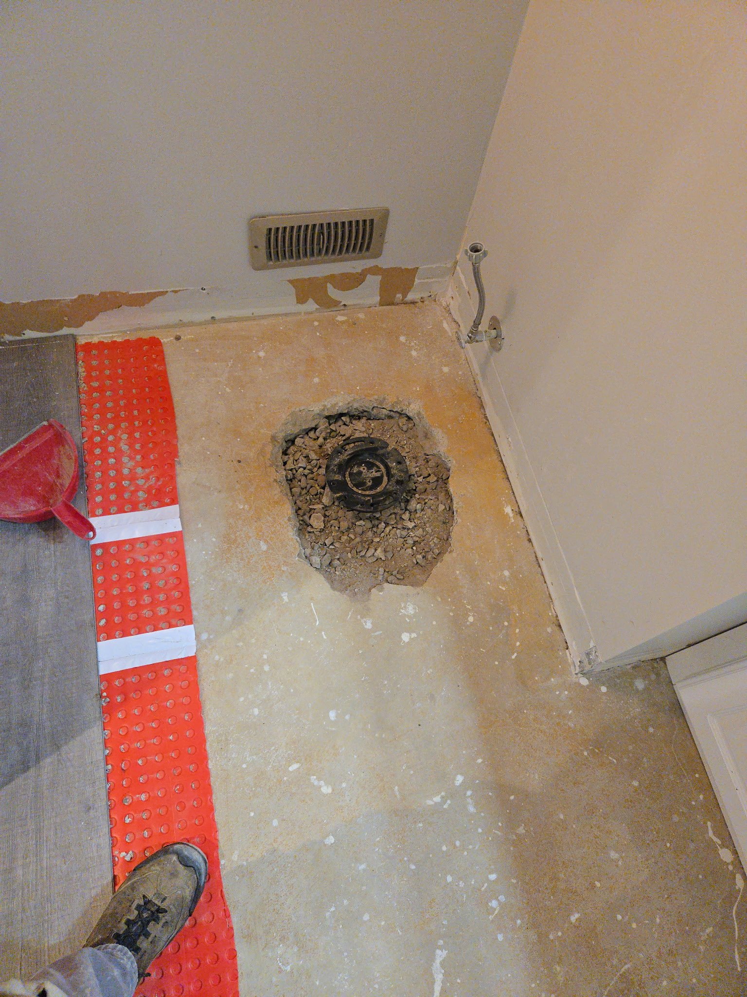 Floor with a hole where a toilet was removed, exposed pipe, vent on the wall, red floor marker, person wearing work boots standing nearby, construction debris scattered around.