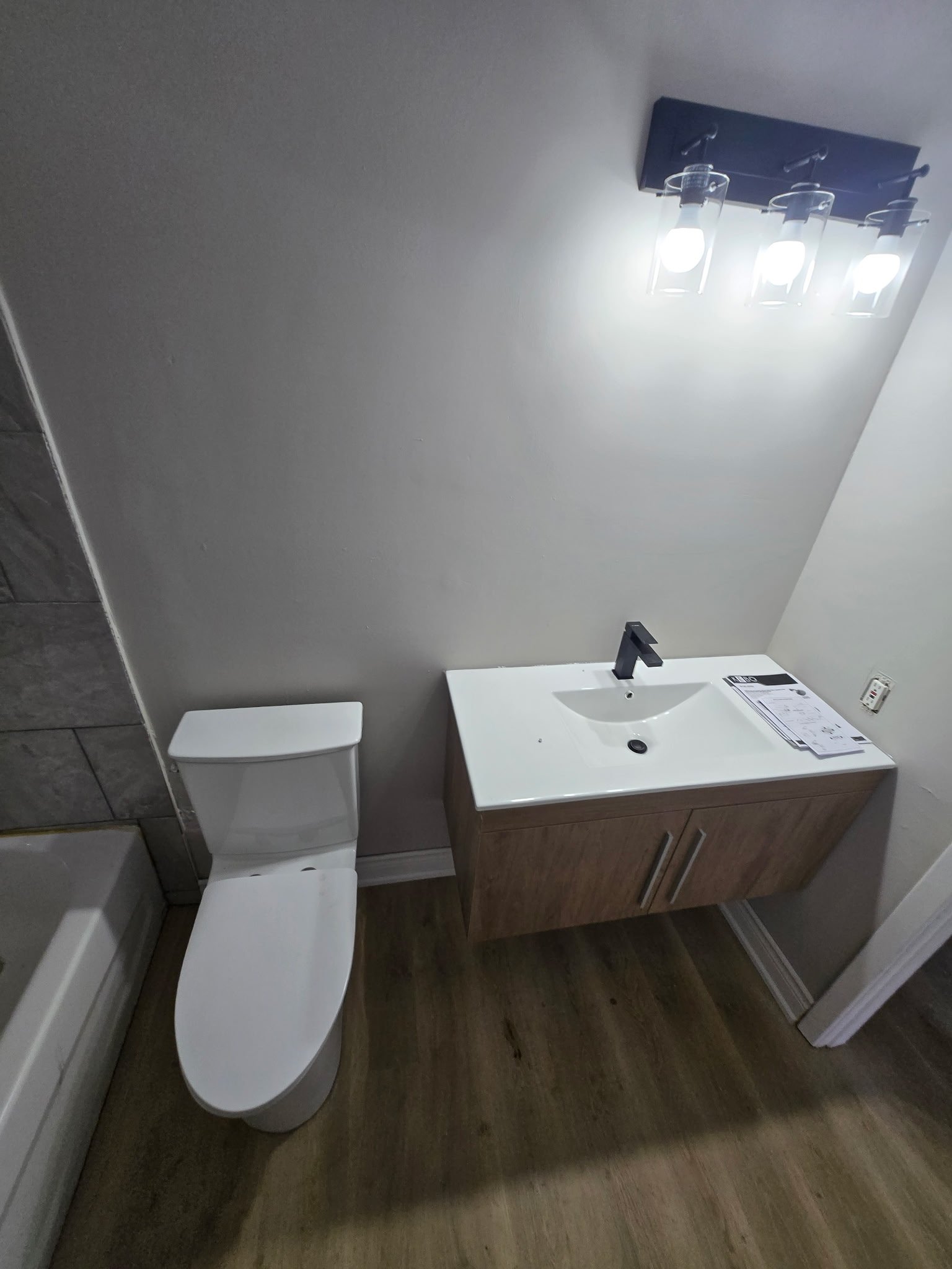 Modern bathroom with a white toilet, a wooden vanity with a white sink, a black faucet, and a mirror above the sink. Bright lighting fixtures on the ceiling, with part of the tiled wall visible on the left.