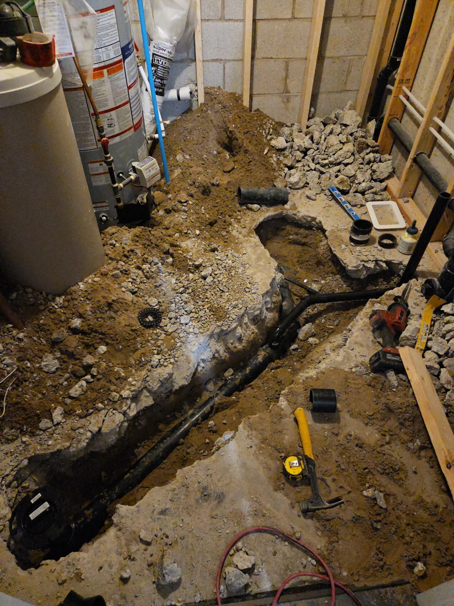 A construction or renovation site with exposed dirt and rocks, plumbing pipes, a water heater, and tools including a level and a measuring tape, ready for plumbing or flooring work.