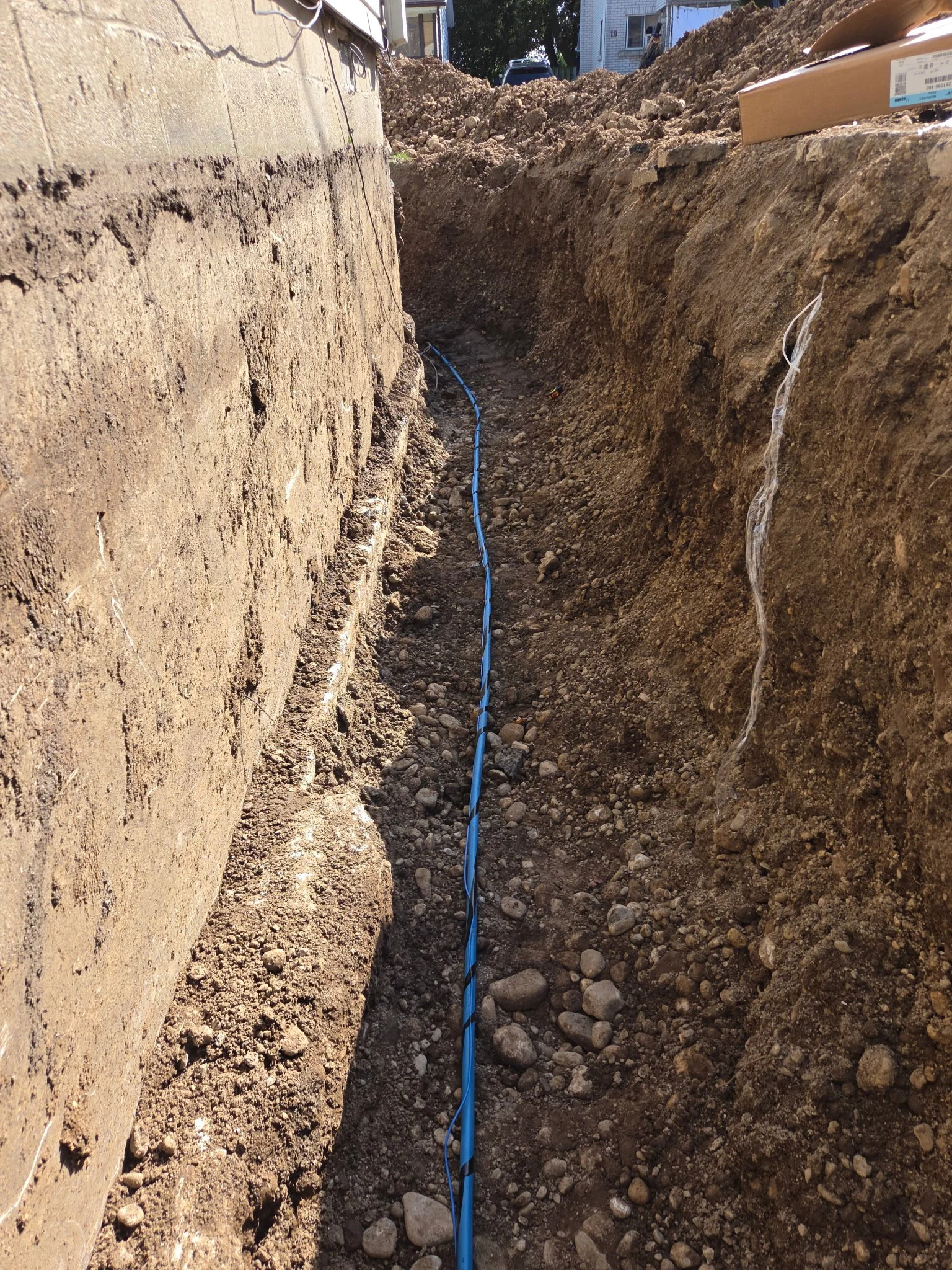 A narrow trench in the ground with a blue underground cable running through it. The trench is next to a wall on the left side, with construction or excavation equipment and houses visible in the background.